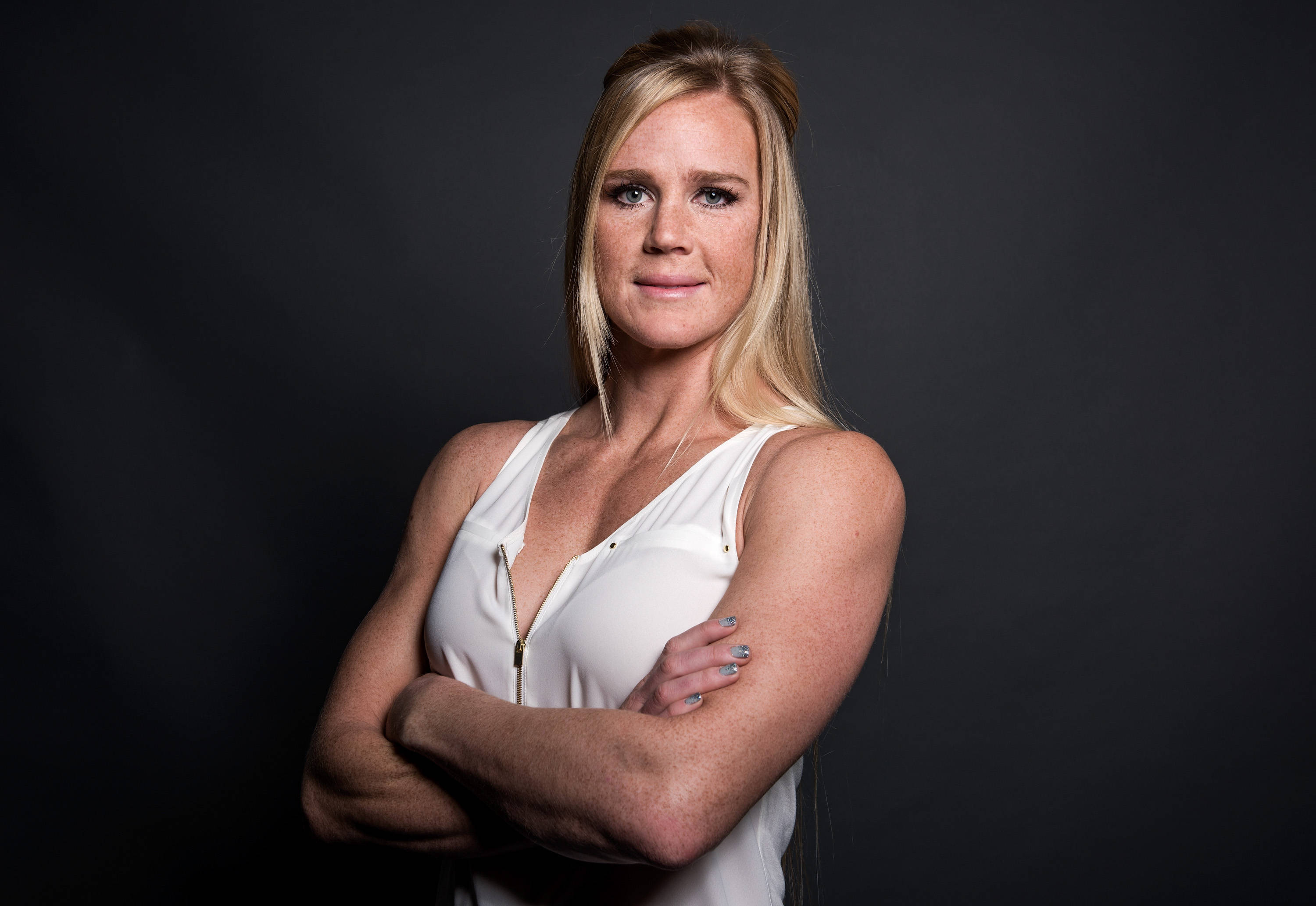 美國MMA選手Holly Holm身穿白色襯衫 壁紙 — 一位自信的金髮女性穿著白色背心，在深色攝影棚背景中交叉雙臂站立。