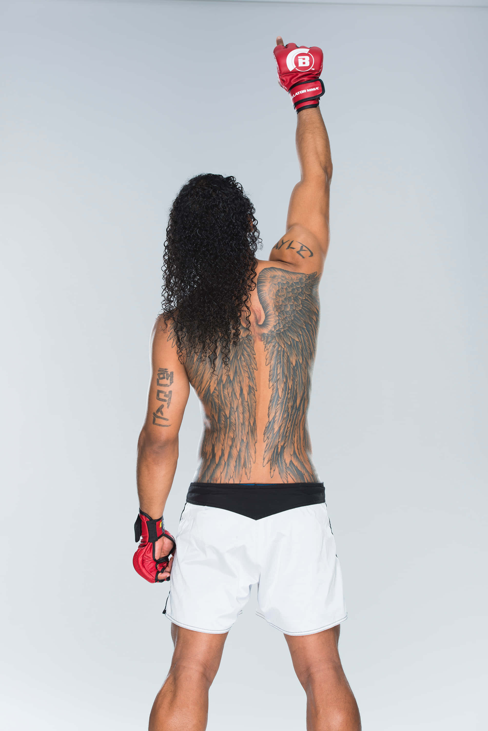 美國MMA選手本·亨德森 Bellator 拍攝照 壁紙 — 一名帶有天使翼紋身的戰士，在純色攝影棚背景下高舉戴手套的拳頭，擺出勝利姿勢。