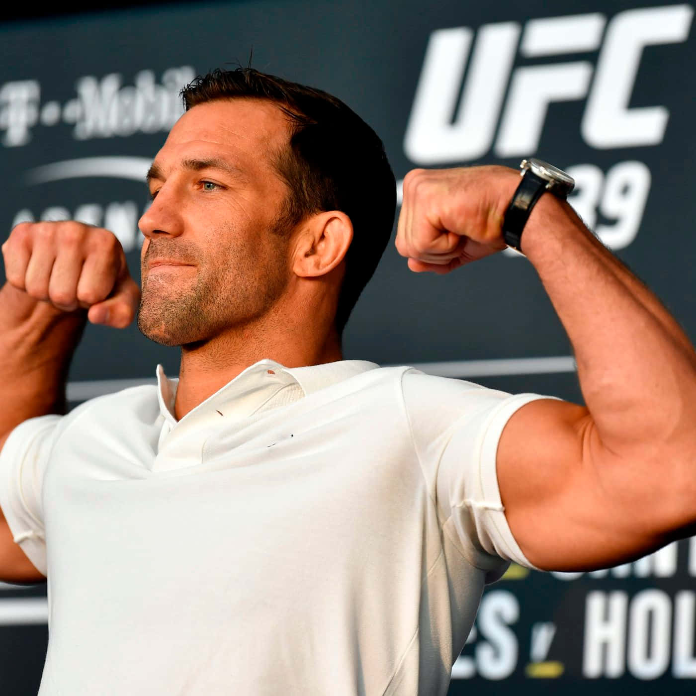美國MMA選手盧克·羅克霍德UFC 239媒體日 壁紙 — 一位肌肉發達的戰士在UFC活動中穿著白色Polo衫，雙臂彎曲，背景帶有品牌標誌。