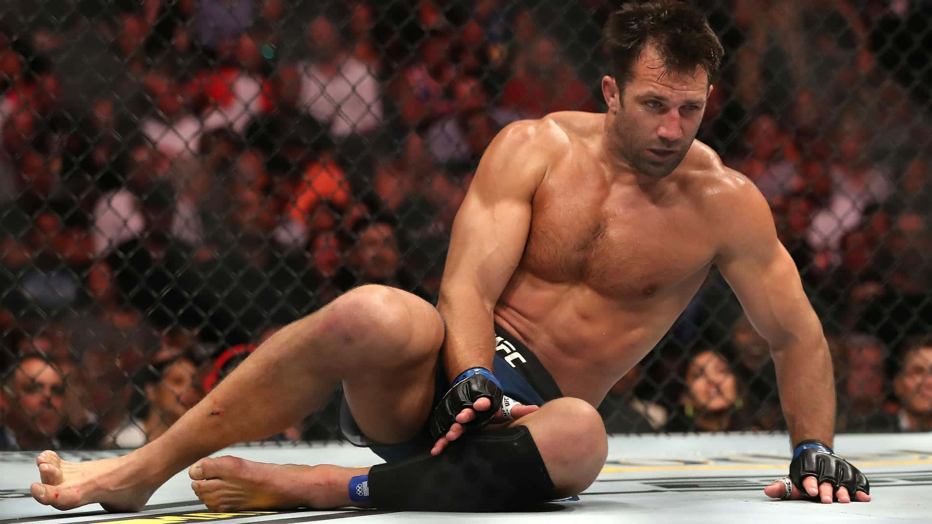 美國UFC戰士盧克·羅克霍德於UFC 239賽事中 壁紙 — 一名肌肉發達的拳手在殘酷的MMA比賽後，疲憊地坐在八角籠地上，籠後的人群模糊不清。