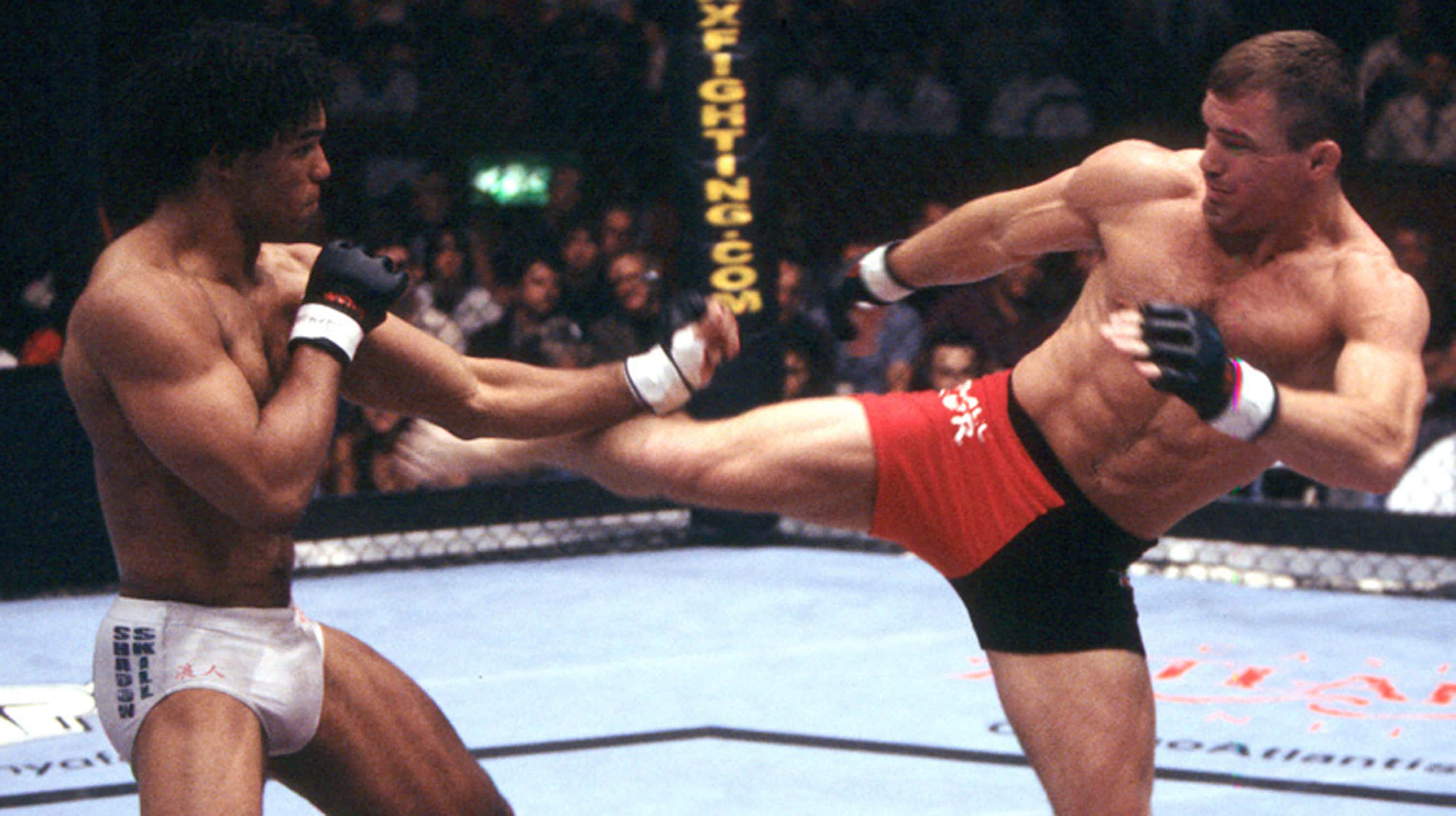 美國UFC戰士肖恩·舍克 壁紙 — 兩名肌肉發達的鬥士在籠中交鋒，一人使出高踢，另一人在明亮的競技場燈光下阻擋。