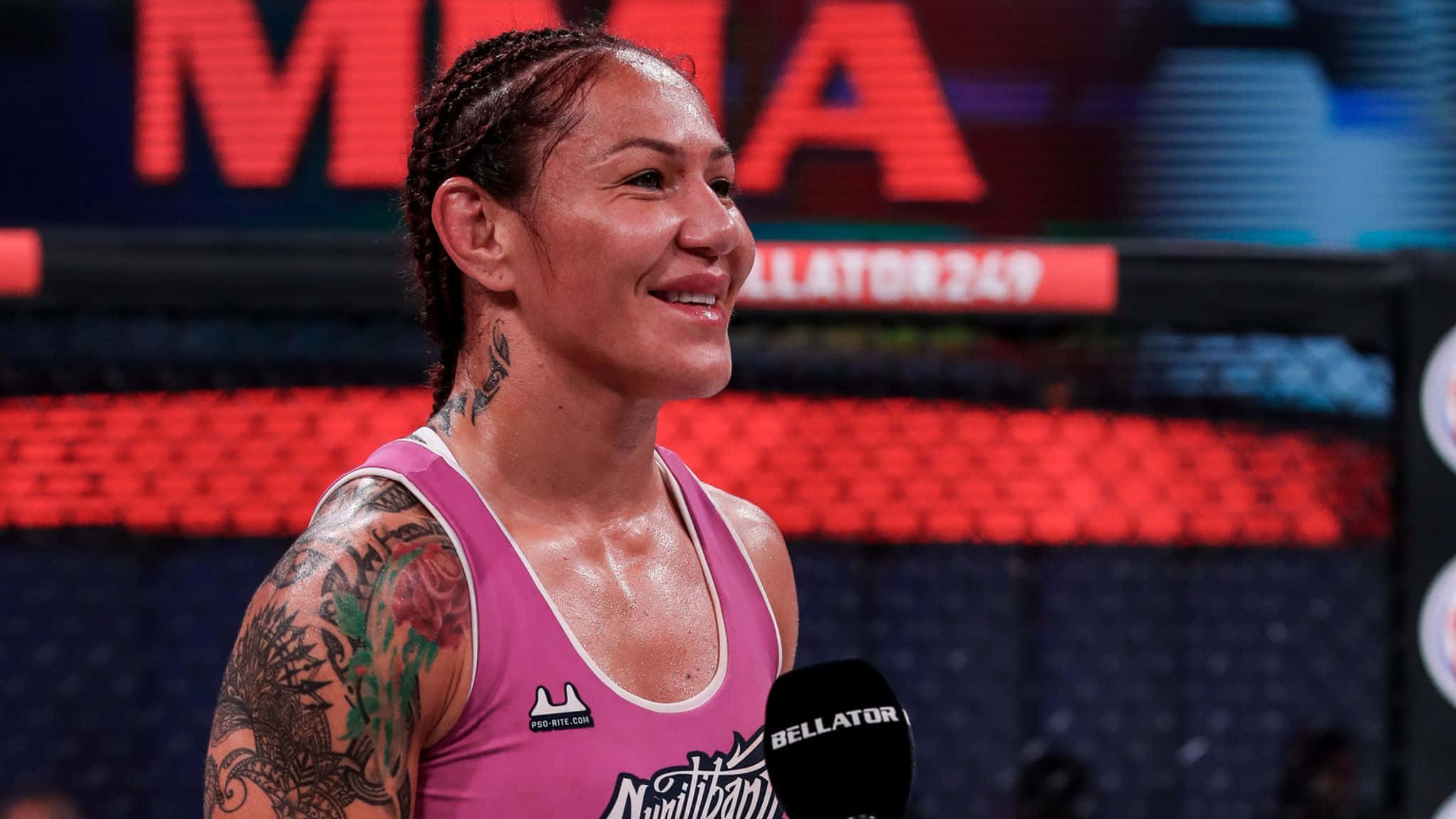 美國UFC選手Cris Cyborg自信微笑照 壁紙 — 一位面帶微笑、有紋身且皮膚濕潤的女性MMA選手，穿著粉紅色連身衣在Bellator籠中接受訪問。