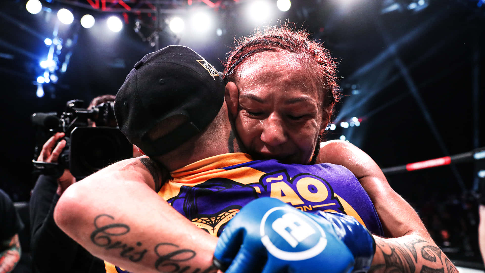 美國UFC選手Cris Cyborg親密擁抱特寫照 壁紙 — 兩名拳手在燈光明亮的競技場上比賽後情感擁抱，周圍閃光燈不斷閃爍。