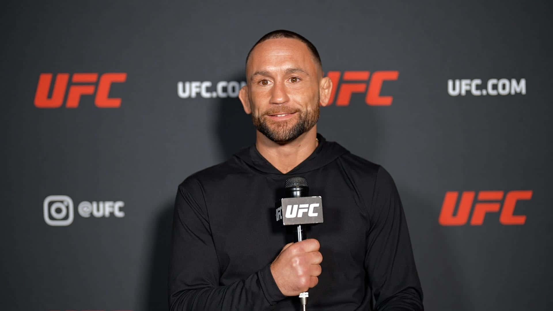 美國UFC選手弗蘭基·愛德加賽前採訪 壁紙 — 一位微笑的戰士穿著黑色連帽衫，在帶有紅色標誌的深灰色背景前握著UFC麥克風。