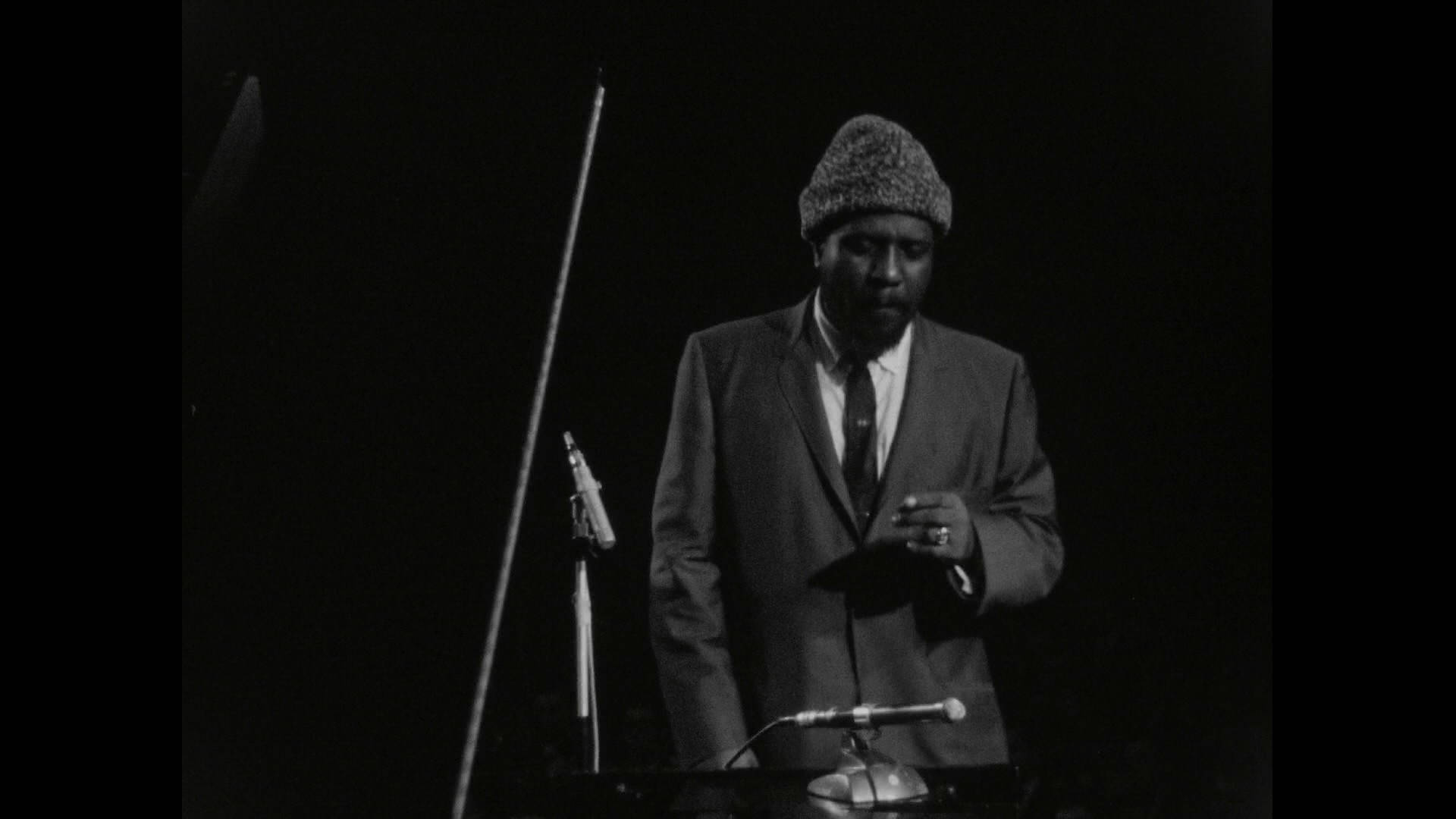 美國作曲家Thelonious Monk 壁紙 — 一名身穿西裝和針織帽的男子在黑白戲劇性舞台燈光下站在鋼琴旁。