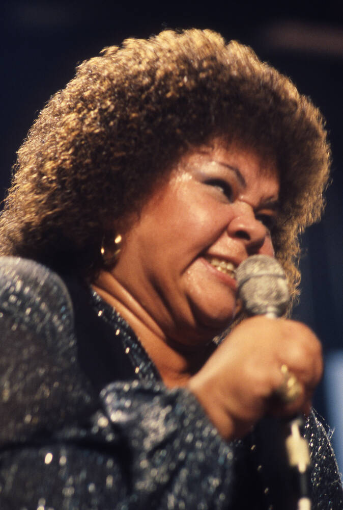 美國偉大歌手 Etta James 壁紙 — 一位快樂的女性，擁有豐盈的捲髮，在舞台燈光下穿著閃亮的藍色服裝對著麥克風唱歌。