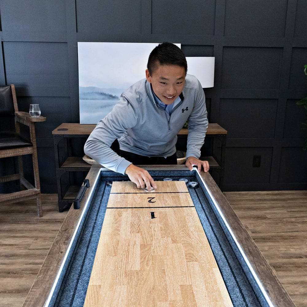 美國傳奇 Kirkwood Shuffleboard 桌 壁紙 — 一名男子微笑著將冰球滑過現代沙狐球桌，房間採用時尚的深色木鑲板。