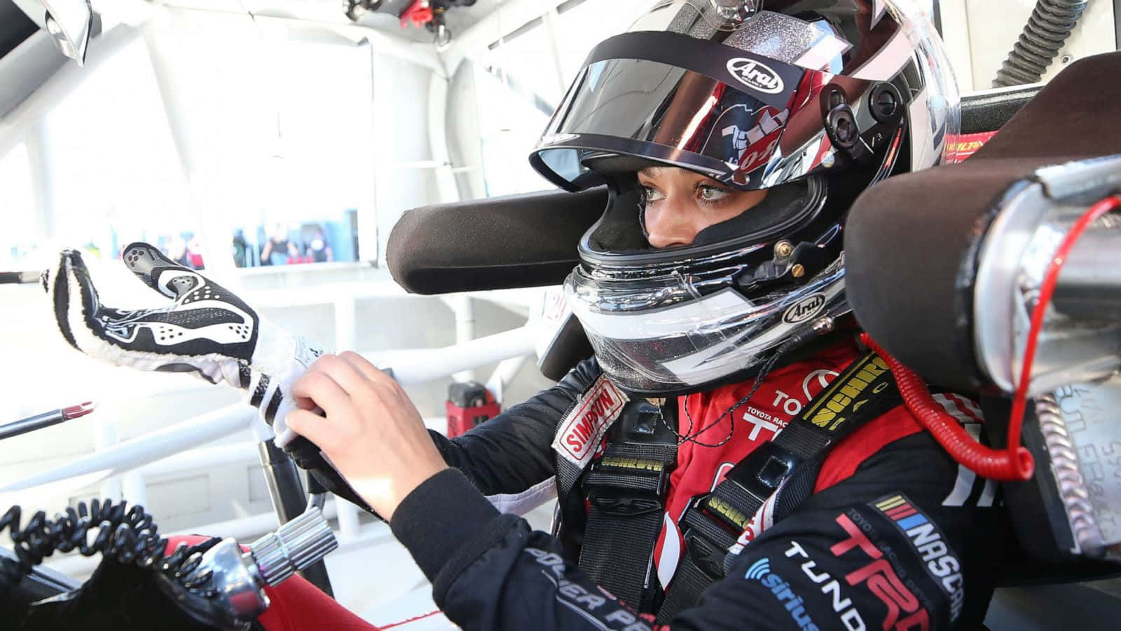 美國傳統——NASCAR賽事 壁紙 — 一位身穿紅黑賽車服和頭盔的賽車手正在賽車駕駛艙內調整手套。
