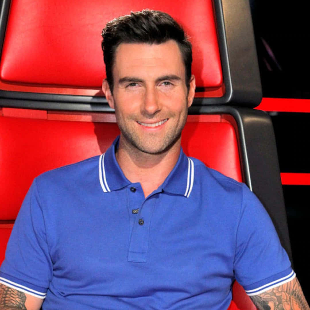 美國創作歌手亞當·萊文 壁紙 — Adam Levine 身穿藍色Polo衫，坐在標誌性的紅色評審椅上，溫暖地微笑。