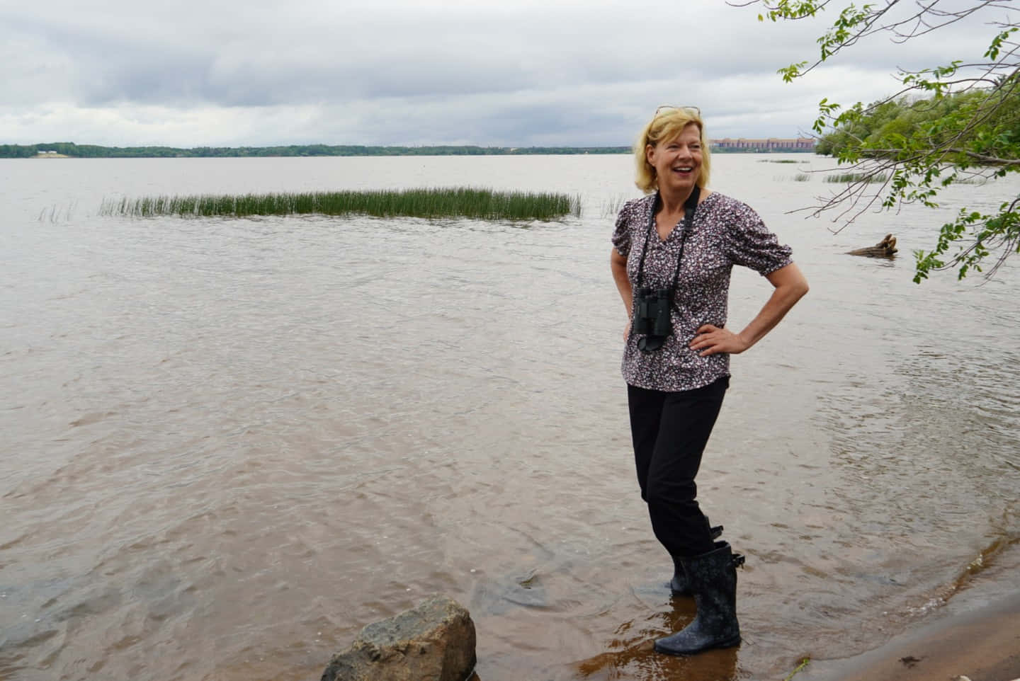 美國參議員 Tammy Baldwin 穿著涉水靴 壁紙 — 一位微笑的女人穿著靴子，站在一個有蘆葦和陰天、水深及膝的泥濘湖中。