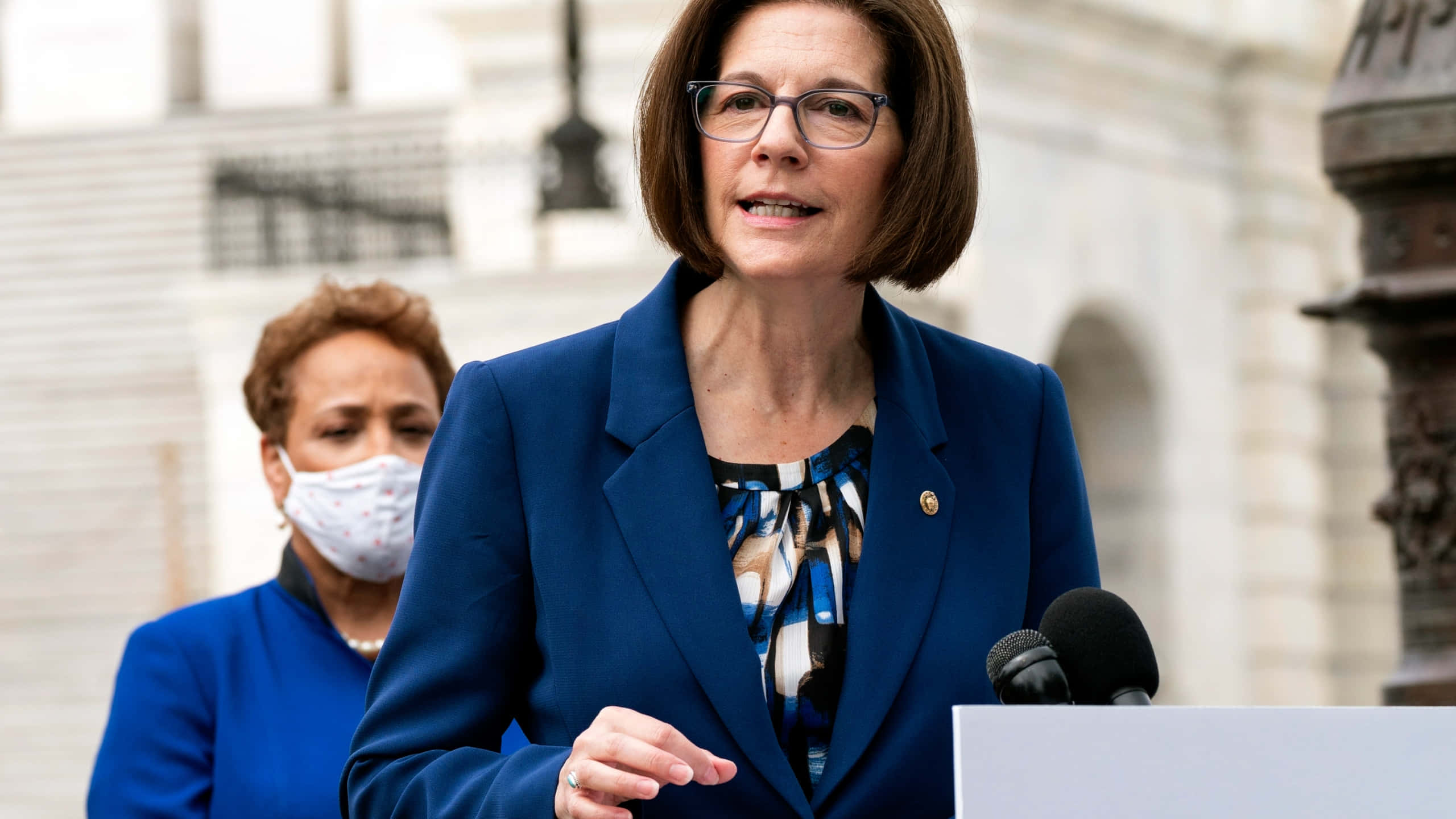 美國參議員卡特琳娜·科爾特斯·馬斯托參與討論 壁紙 — 一位身穿藍色西裝外套的女性在戶外講台上發言，背後是一座古典建築，另一位戴著口罩的女性站在她身後。