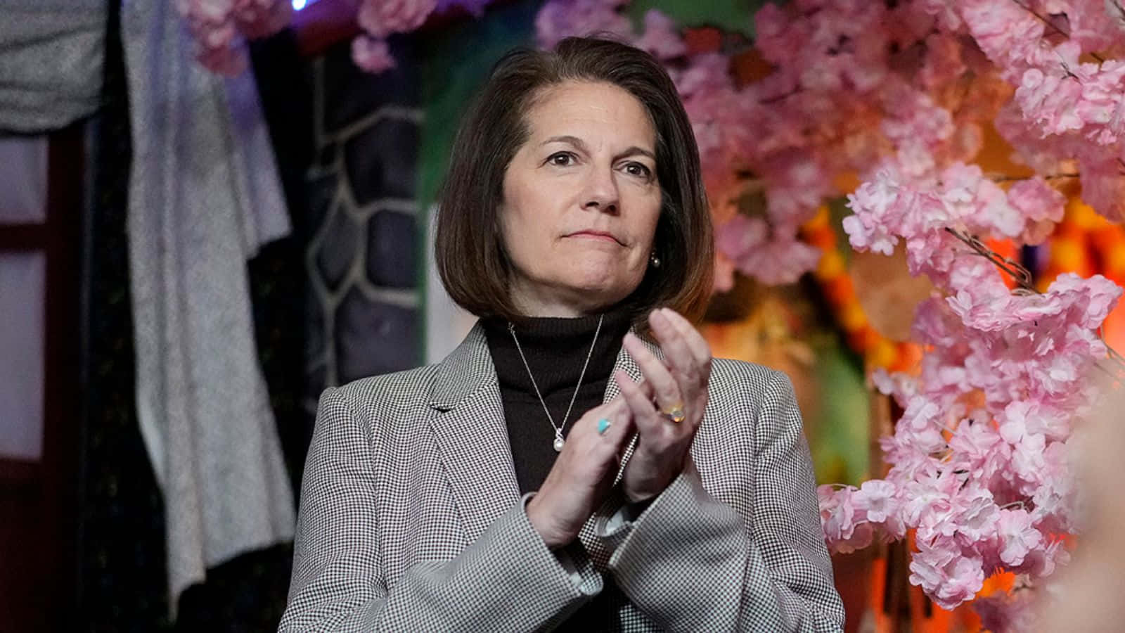 美國參議員卡特里娜·科爾特斯·馬斯托，內華達州在華盛頓的聲音 壁紙 — 一位穿著灰色格紋西裝外套的女性，在充滿活力的粉紅色櫻花和溫暖發光的燈光背景下拍手。