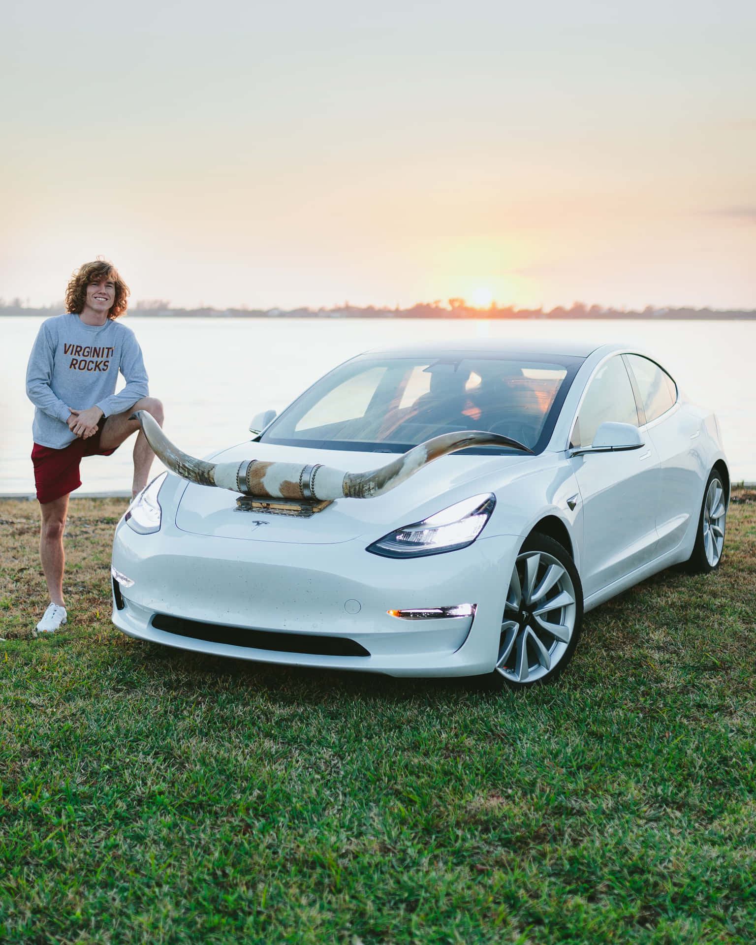 年輕男子在日落湖邊與裝有公牛角的白色Tesla汽車合影。