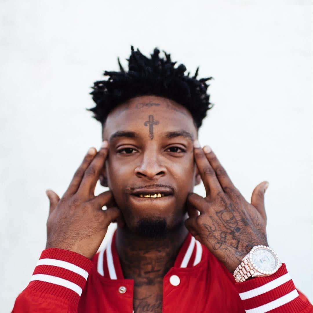 美國嘻哈藝人21 Savage，以陷阱音樂與強烈歌詞聞名。 壁紙 — 一名額頭有十字紋身、雙手佈滿紋身的男子，在純白背景前將手指按在太陽穴上。