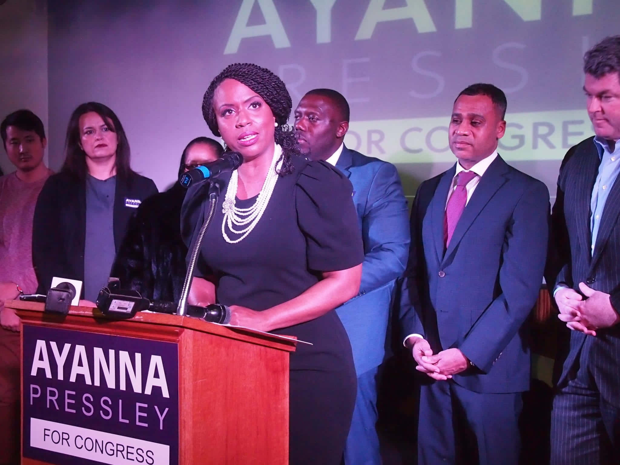 美國國會議員艾安娜·普雷斯利與同事進行深刻對話 壁紙 — Ayanna Pressley 在舞台燈光下於競選講台上發言，支持者站在她身後。