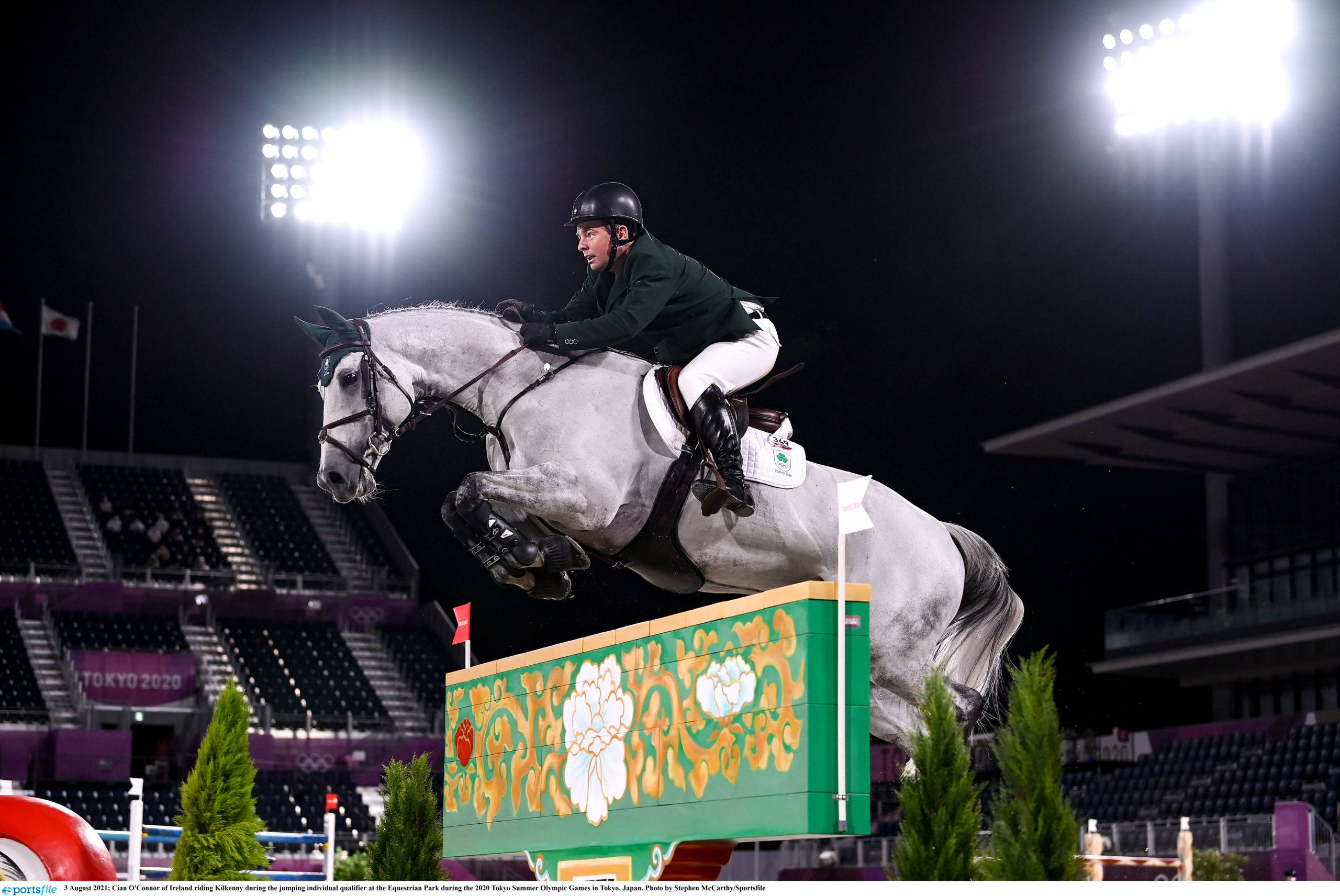 美國奧運女騎手勞拉·克勞特參加冬季馬術節 壁紙 — 一名身穿綠色和白色衣服的騎手在夜間明亮的體育場燈光下引導一匹灰色馬匹跳過色彩繽紛的障礙物。