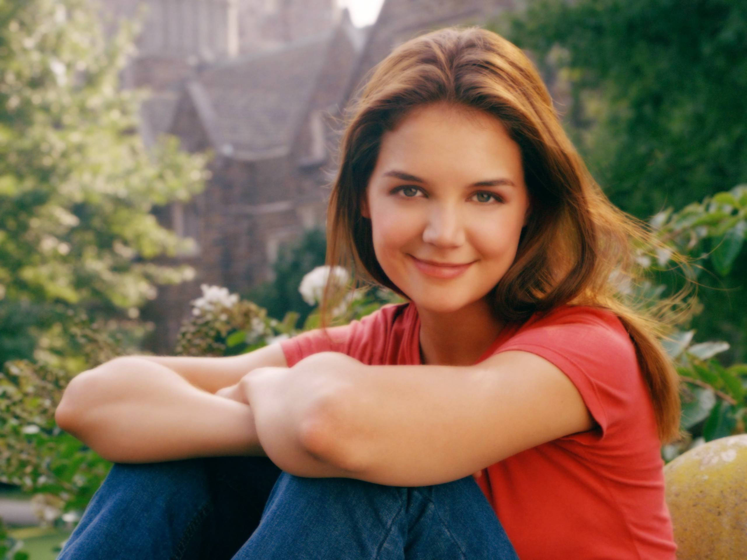 美國女演員凱蒂·霍爾姆斯 飾演《 Dawson's Creek 》中的喬伊·波特 壁紙 — 一位身穿紅色襯衫的年輕女性在戶外微笑，身後有綠植和石頭建築。