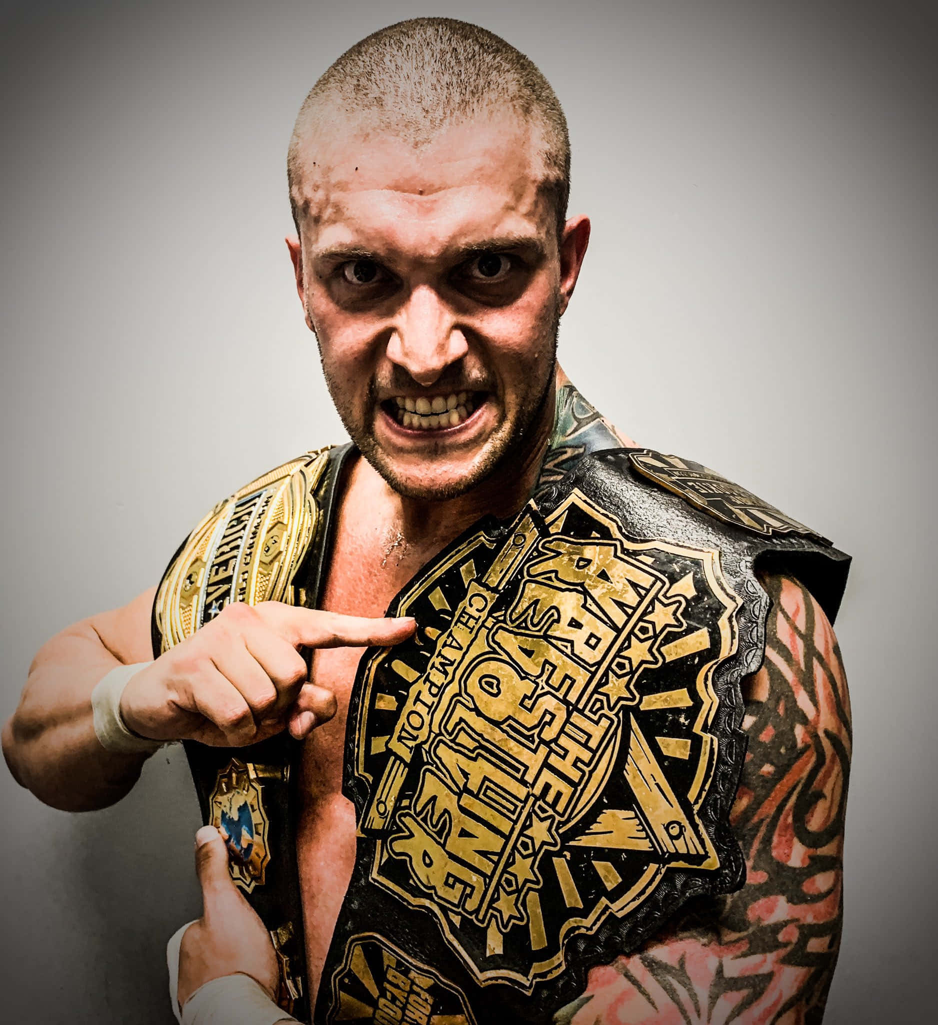 美國摔角手 Killer Kross 展示 Wrestling Revolver 擊敗腰帶 壁紙 — 一個兇猛的光頭紋身摔角手，在純灰色背景前指向他的冠軍腰帶。