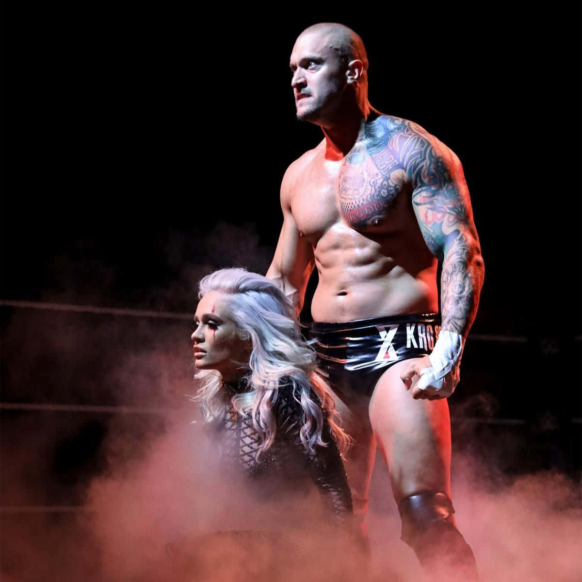 美國摔角手 Killer Kross 與妻子 Scarlett 在 WWE Basic 120 的側面視角 壁紙 — 一位有紋身的摔角手和一位金髮女子站在煙霧瀰漫的擂台上，在戲劇性的紅燈光下。