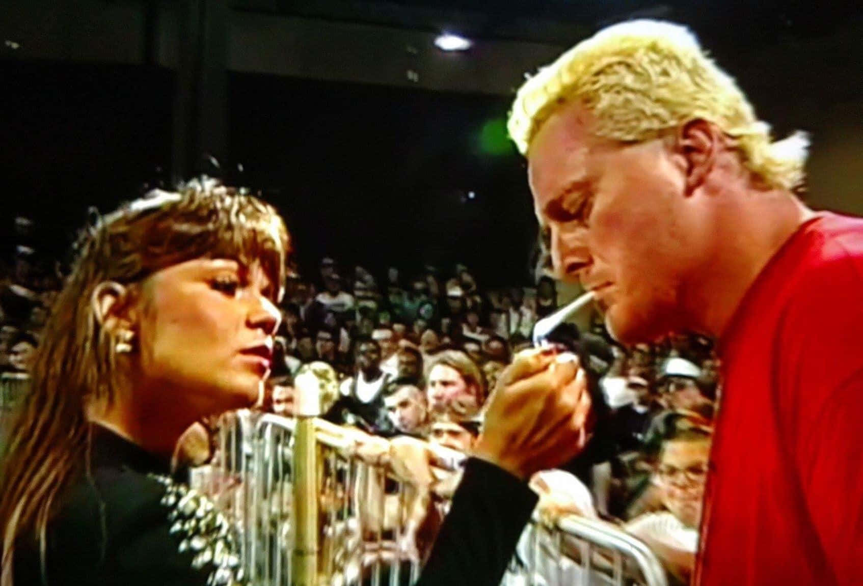 美國摔角手 The Sandman 與 Nancy Benoit 吸煙 壁紙 — 一名金髮男子身穿紅色襯衫，在擠滿人的活動中俯身越過欄杆，為一名身穿黑色服裝的女子點煙。