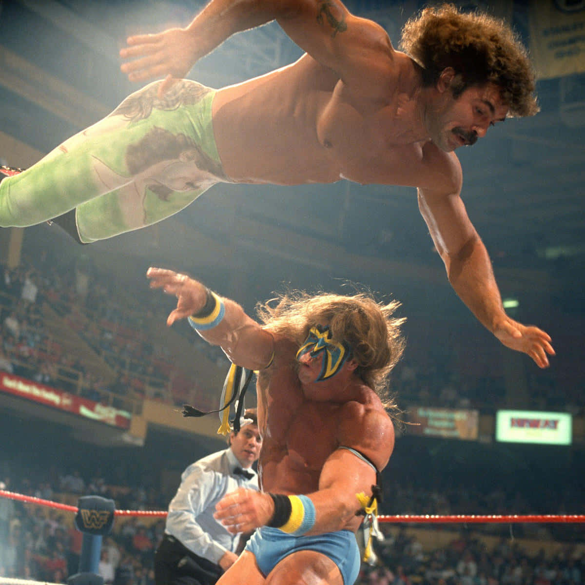 美國摔角手 Ultimate Warrior 與 Rick Rude 1989 年 Summerslam 壁紙 — 一個肌肉發達的摔角手在燈光明亮的擂台上，向對手飛身撲去，背景是歡呼的人群。