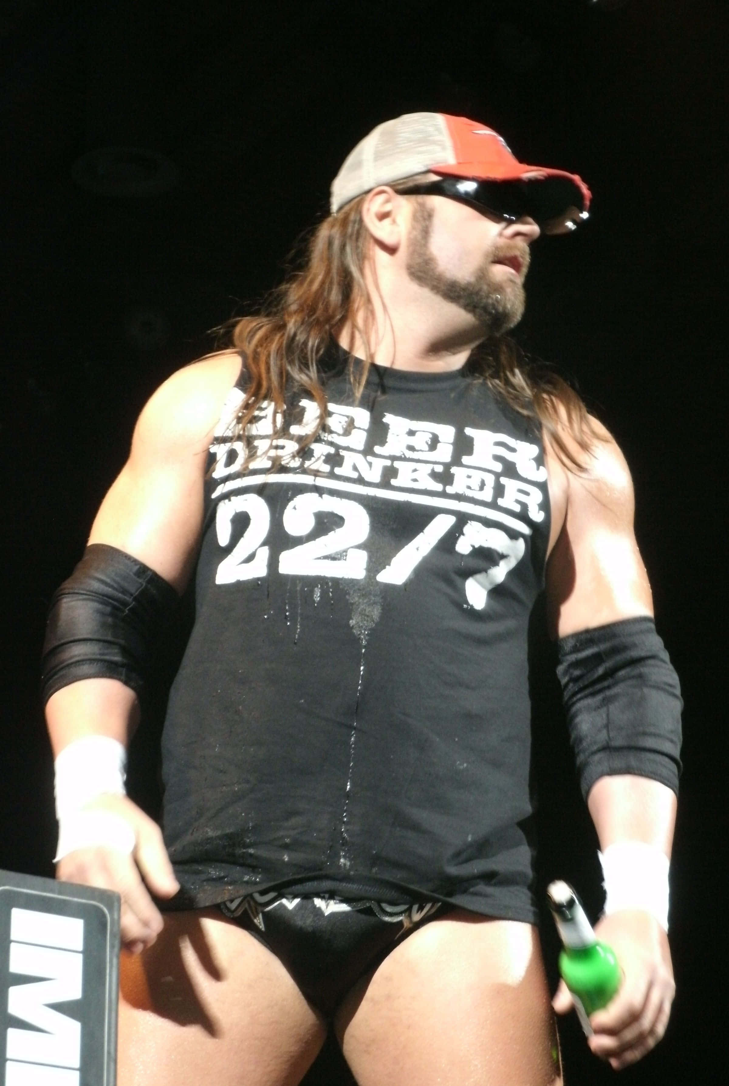美國摔角手Cowboy James Storm 2012年TNA賽事 壁紙 — 一位肌肉發達的摔角手身穿黑色背心和紅色帽子，在明亮的舞台燈光下拿著一個綠色瓶子。