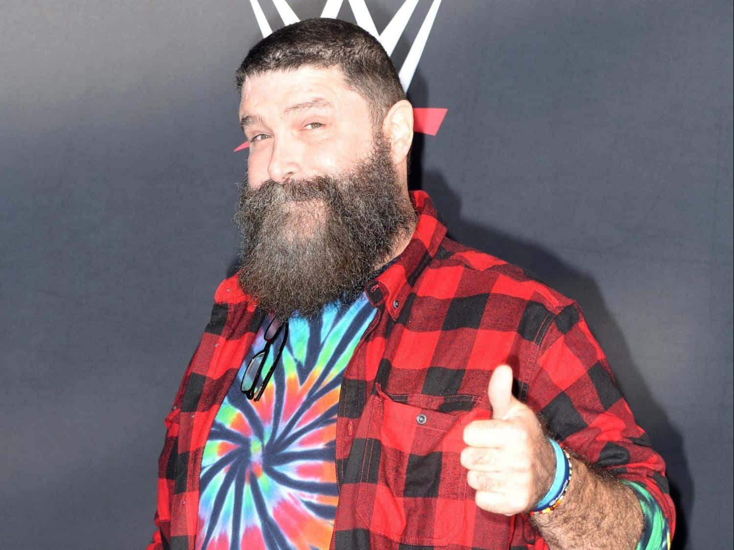 美國摔角手米克·福萊出席WWE 20週年慶典 壁紙 — 一位留鬍鬚的男人穿著紅色格仔恤衫內搭紮染T恤，在黑暗背景前豎起大拇指，背景有WWE標誌。