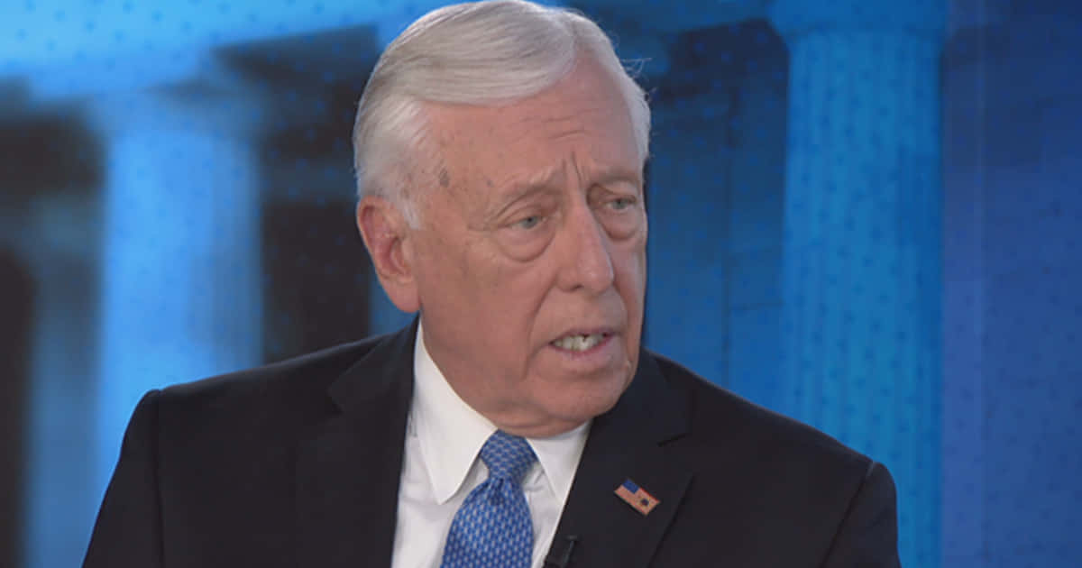 美國政治人物 Steny Hoyer 接受訪問 壁紙 — 一位身穿深色西裝和藍色領帶的年長男性，在模糊的藍色背景前說話的特寫。