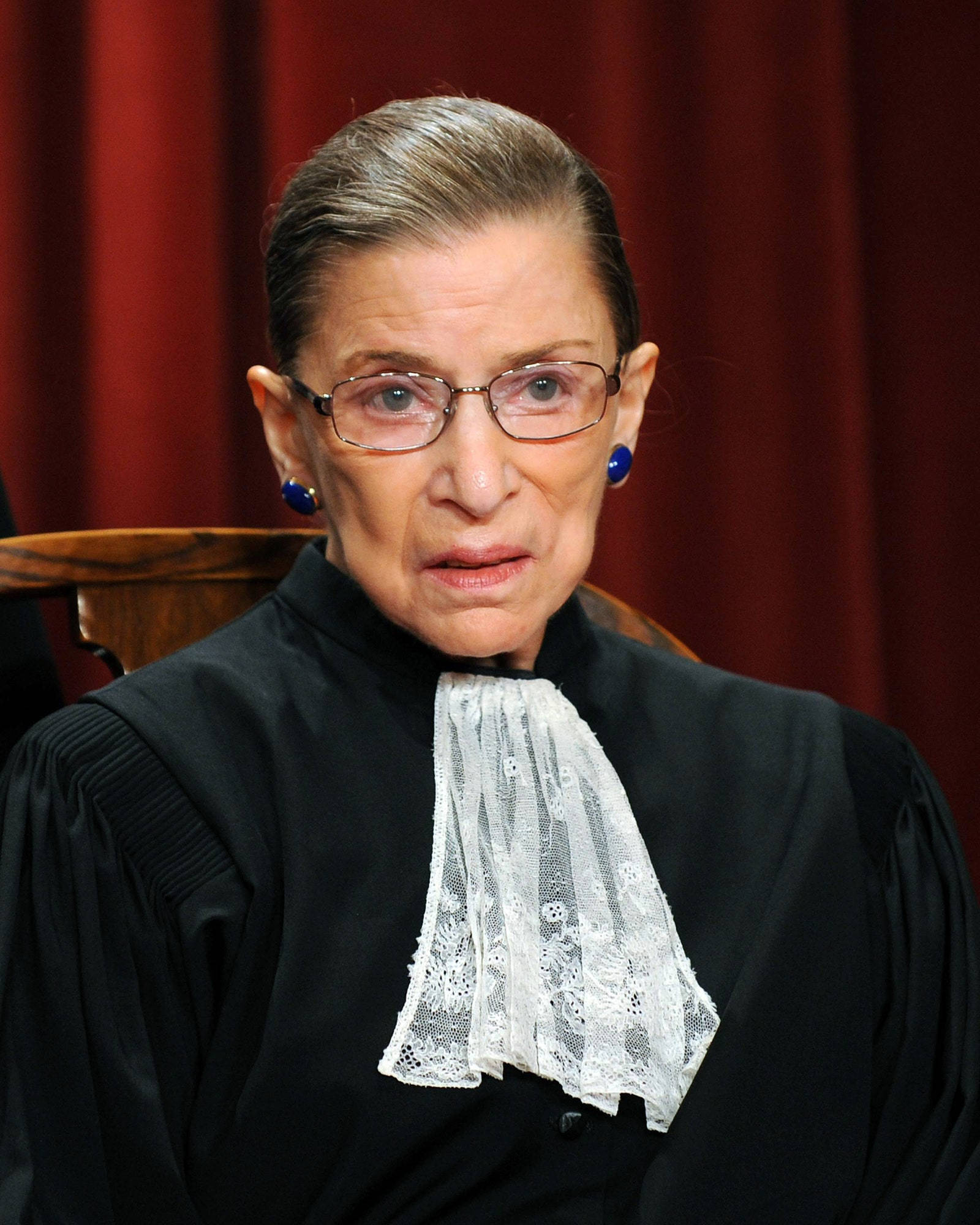 美國最高法院大法官露絲·巴德·金斯伯格 壁紙 — Ruth Bader Ginsburg 身穿黑色法官袍、白色蕾絲領飾，坐在紅色帷幕前的肖像。