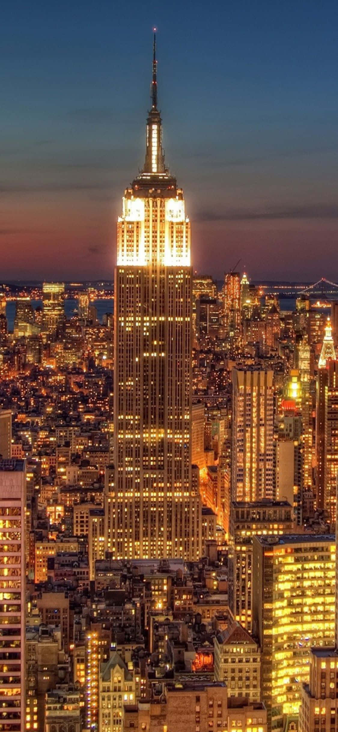 美國標誌性城市紐約的迷人風景 壁紙 — 帝國大廈在暮色天空中閃耀金色光芒，紐約市的燈光在下方閃爍。