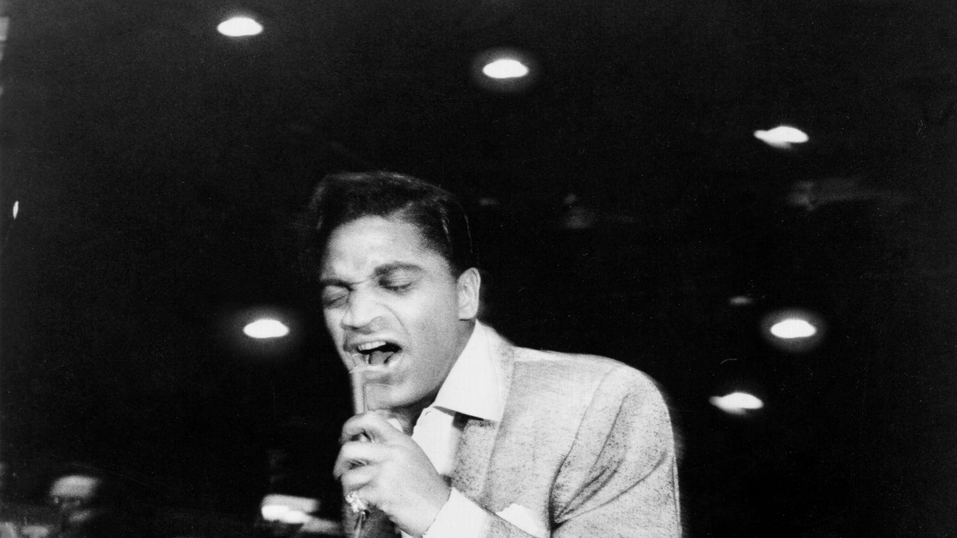 美國歌手Jackie Wilson - 摇滾樂 壁紙 — 一張黑白照片捕捉到一位充滿激情的男歌手在舞台燈光下拿著麥克風表演的瞬間。