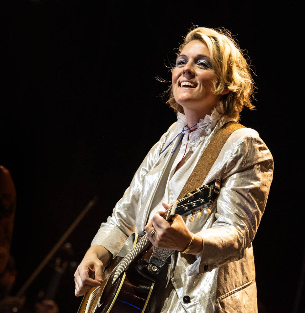 美國歌手布蘭迪·卡莉 壁紙 — 一位微笑的金髮女性穿著閃亮的銀色外套，在黑色背景的舞台燈光下彈奏木結他。