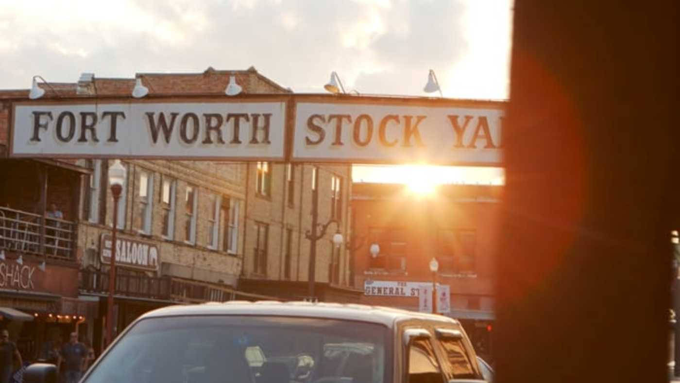 美國沃斯堡市中心 壁紙 — 夕陽在歷史悠久的Fort Worth Stockyards標誌後方發光，一輛汽車駛過復古建築。