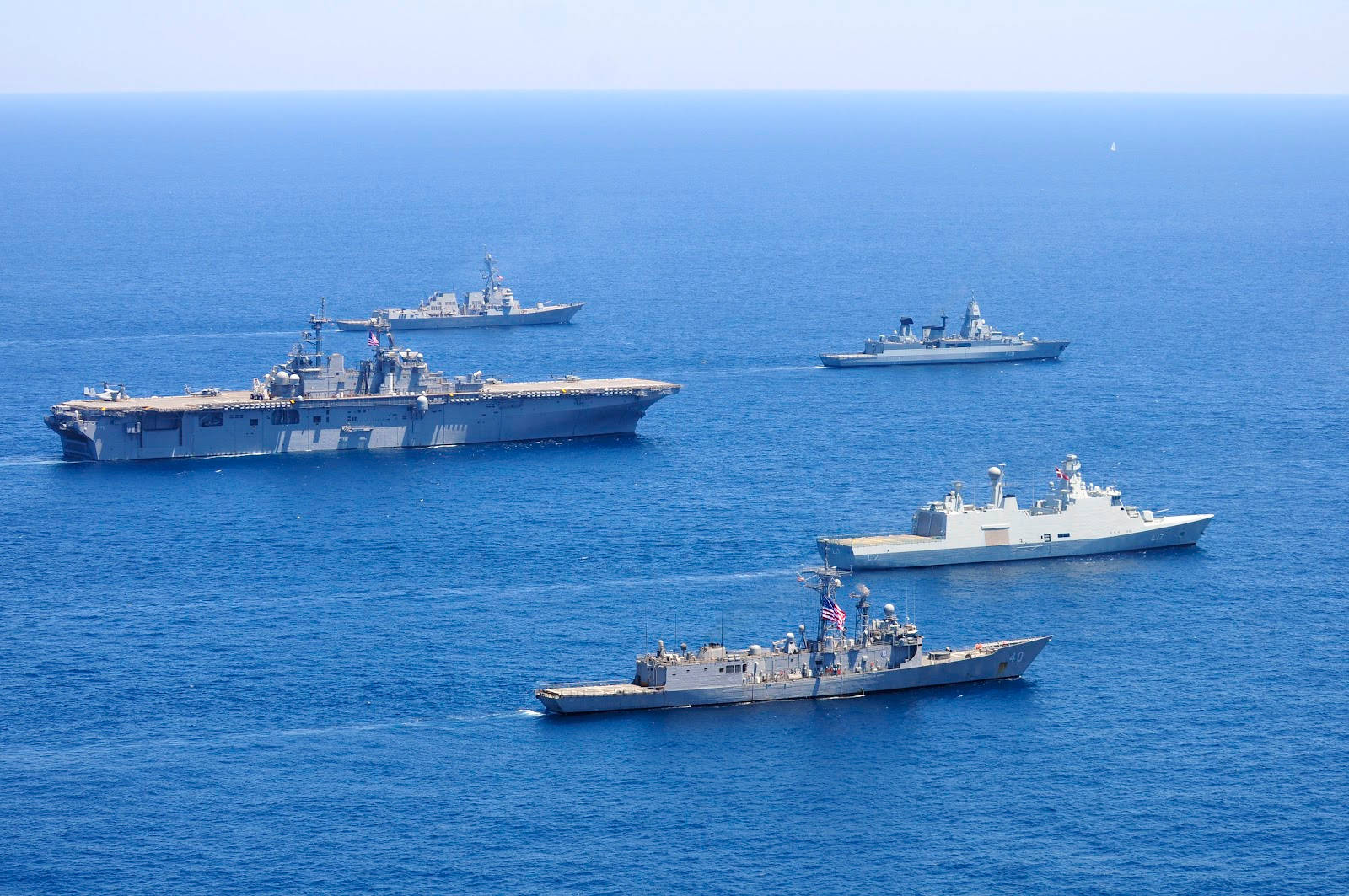 美國海軍兩棲攻擊艦與其他艦艇同框 壁紙 — 四艘海軍戰艦（包括一艘航空母艦）在晴朗的天空下，以編隊形式航行在廣闊的深藍色海洋上。