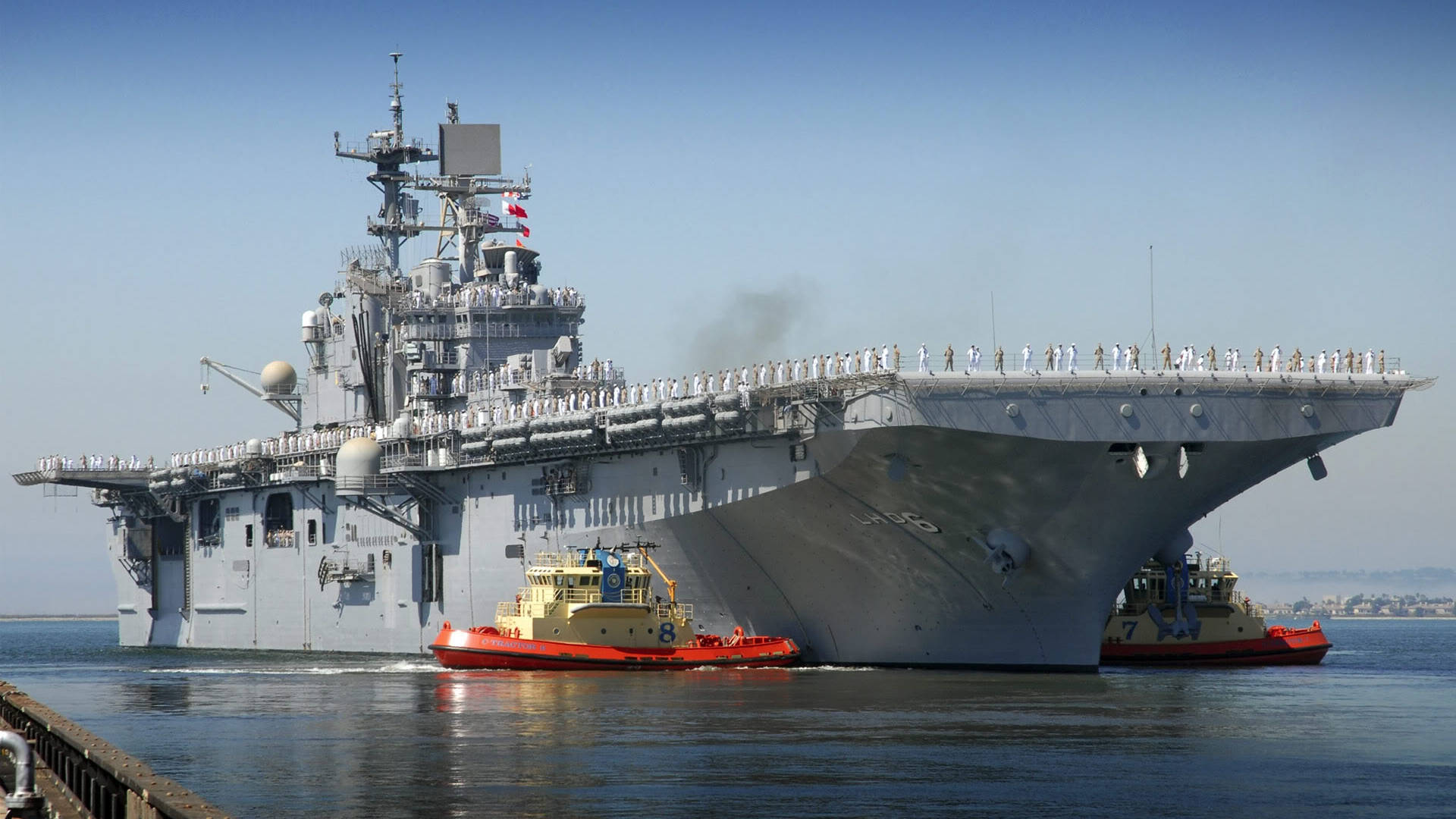 美國海軍兩棲攻擊艦被拖曳 壁紙 — 一艘巨大的灰色航空母艦，甲板上有船員，在晴朗的天空下，由兩艘紅色和黃色的拖船引導，穿過平靜的藍色水域。