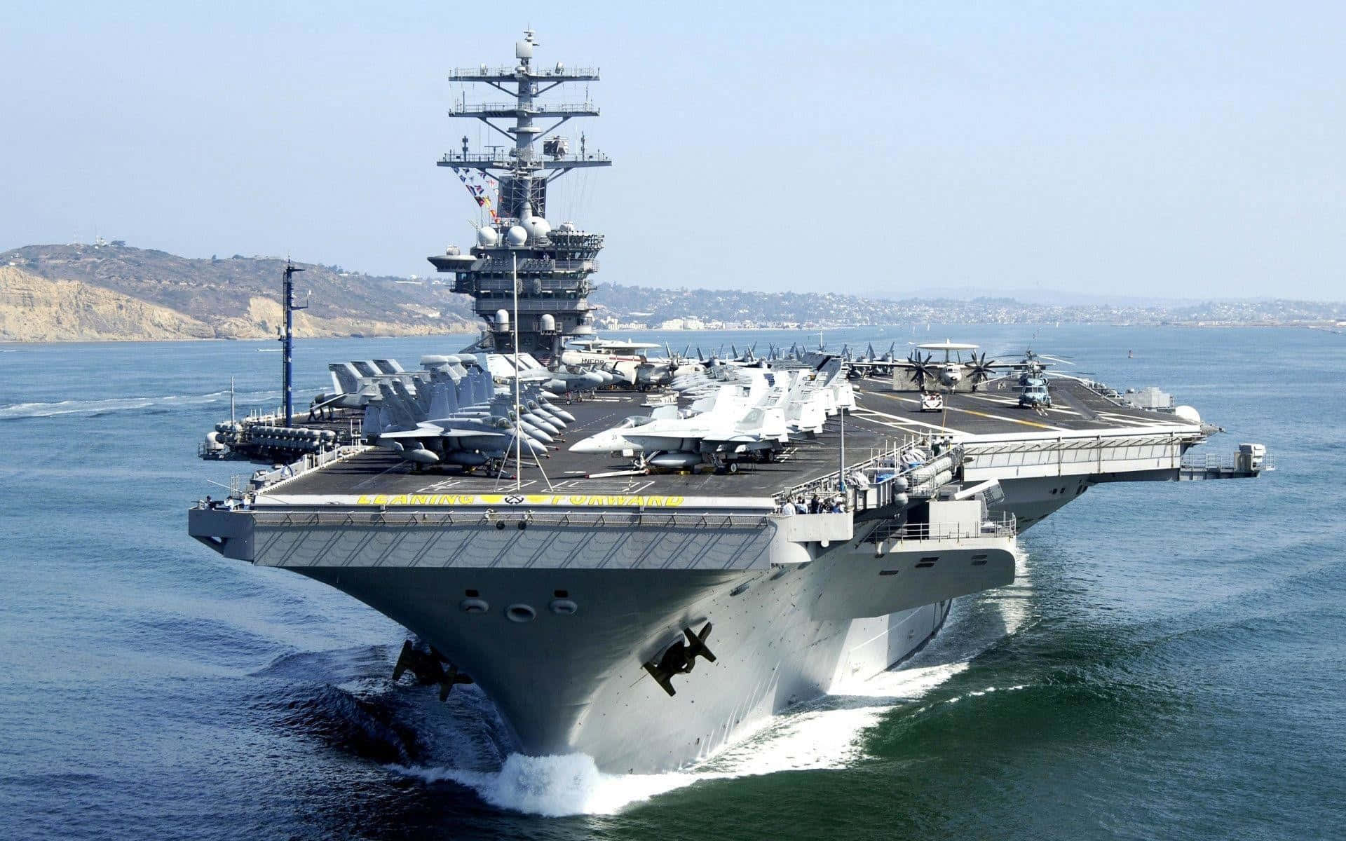 美國海軍堅強挺立！ 壁紙 — 一艘巨大的灰色航空母艦在藍色海洋中航行，甲板上排列著戰鬥機，天空晴朗。