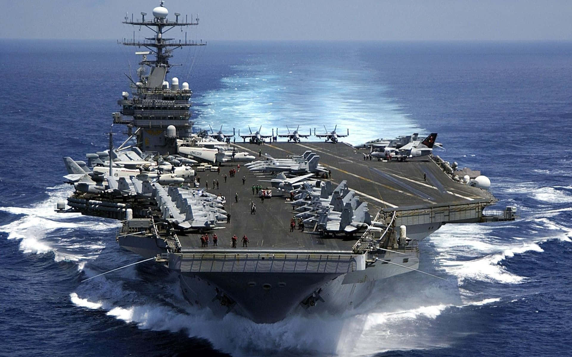 美國海軍的驕傲 壁紙 — 一艘巨大的航空母艦在深藍色的海浪中破浪前行，戰鬥機整齊排列在甲板上，天空晴朗。