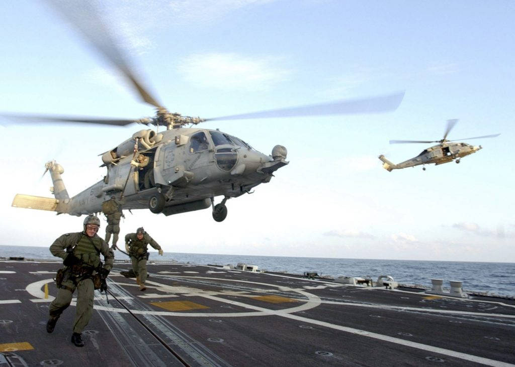 美國海軍直升機降落航空母艦 壁紙 — 兩架軍用直升機在航空母艦甲板上空盤旋，水兵們在晴朗的藍天下向船邊衝刺。