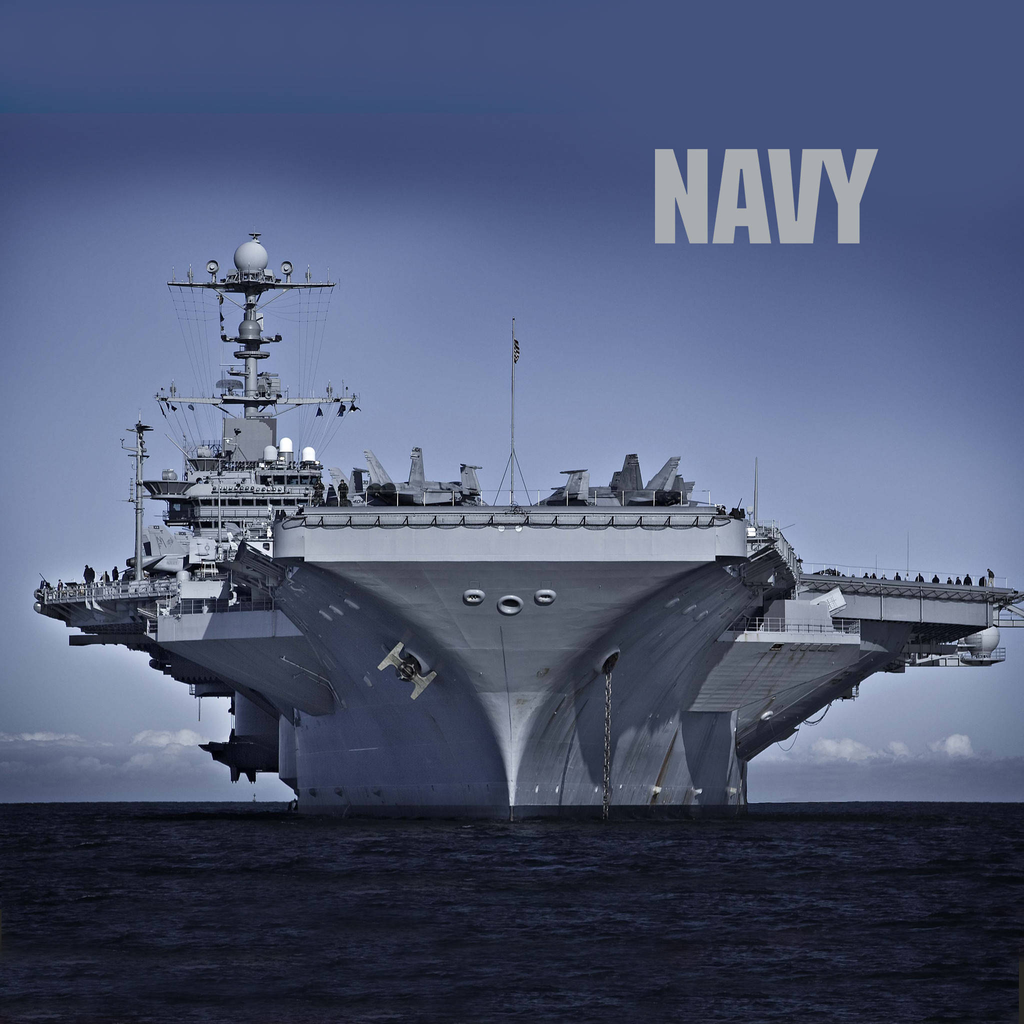 美國海軍航空母艦向前航行 壁紙 — 一艘巨大的灰色航空母艦在晴朗的藍天下主宰海洋，右上角有「NAVY」文字。