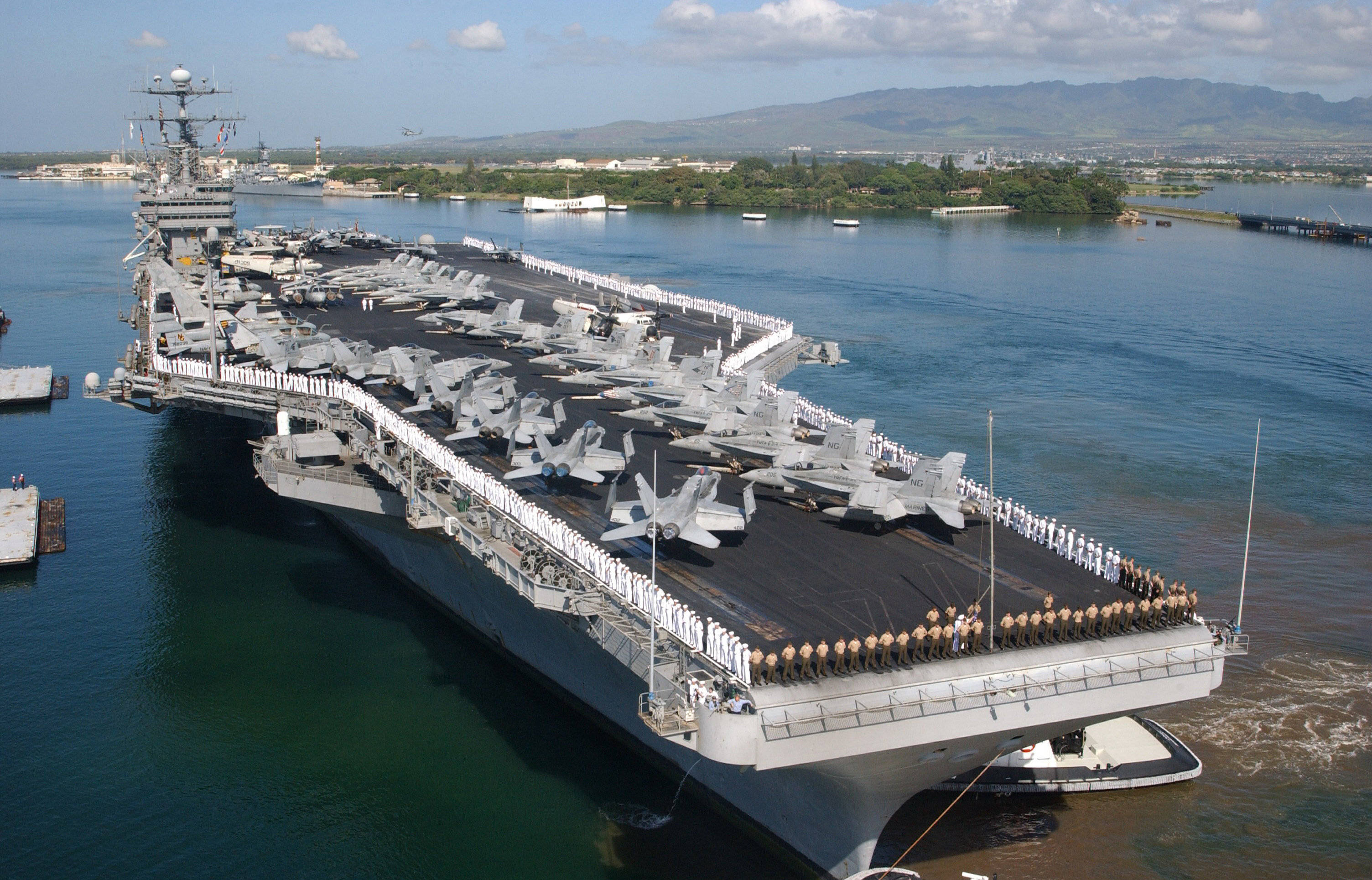 美國海軍航空母艦滿載戰機 壁紙 — 一艘巨大的灰色航空母艦停泊在平靜的藍色水域中，甲板上排列著戰鬥機，水兵們沿著欄杆立正站著。