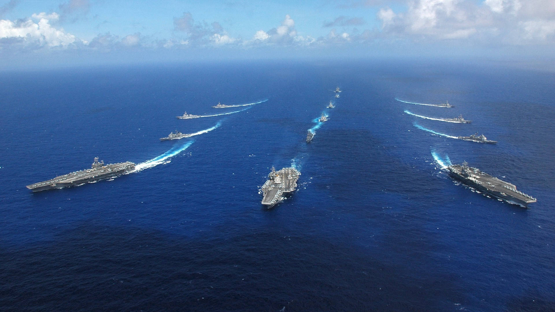 美國海軍航空母艦編隊航行 壁紙 — 從空中俯瞰大規模海軍艦隊，包括航空母艦和驅逐艦，在部分多雲的天空下穿越深藍色海洋。