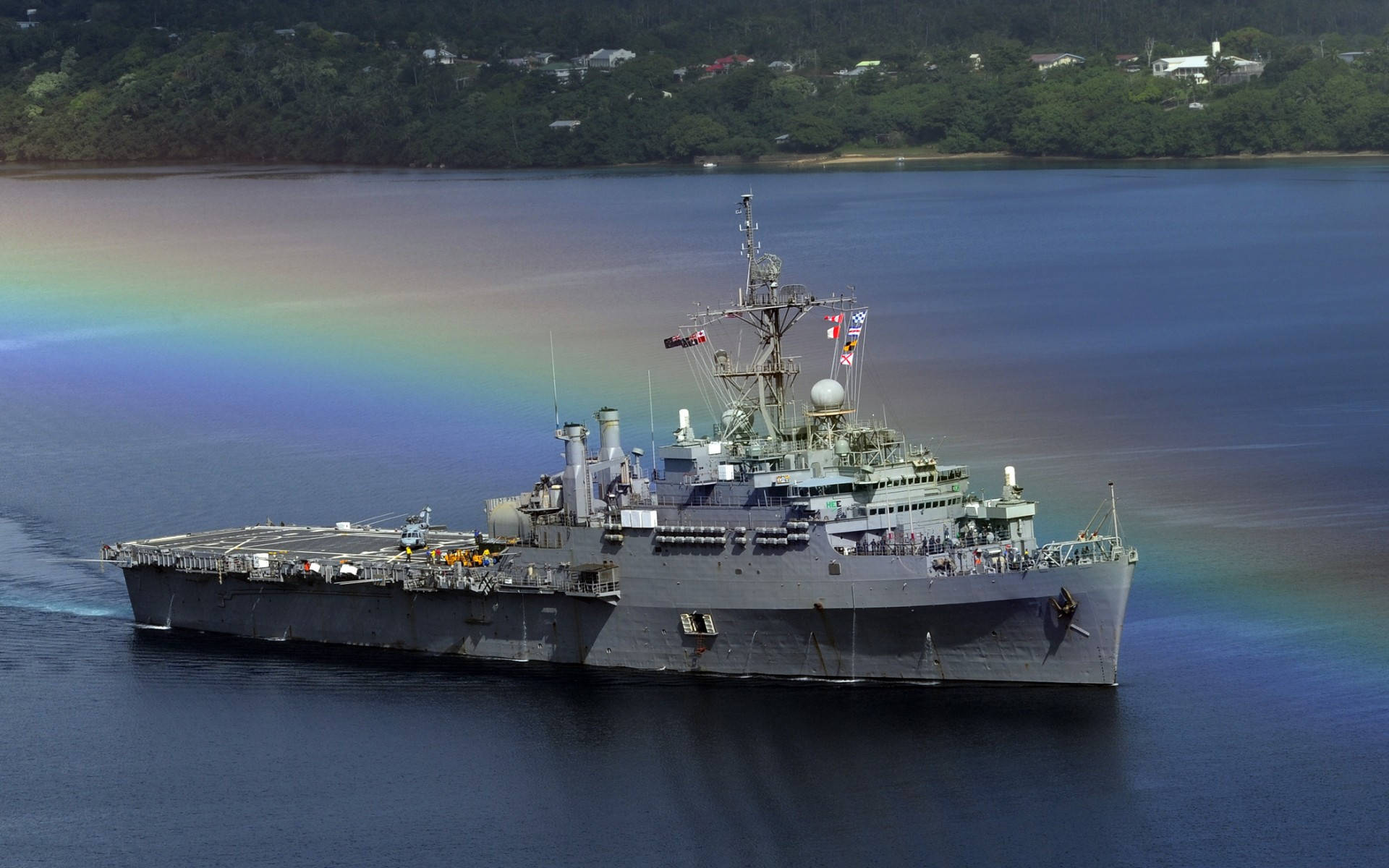 美國海軍艦艇背後彩虹 壁紙 — 一艘灰色軍艦在平靜的藍色海面上航行，背後有一道鮮豔的彩虹在樹木繁茂的海岸線附近拱起。