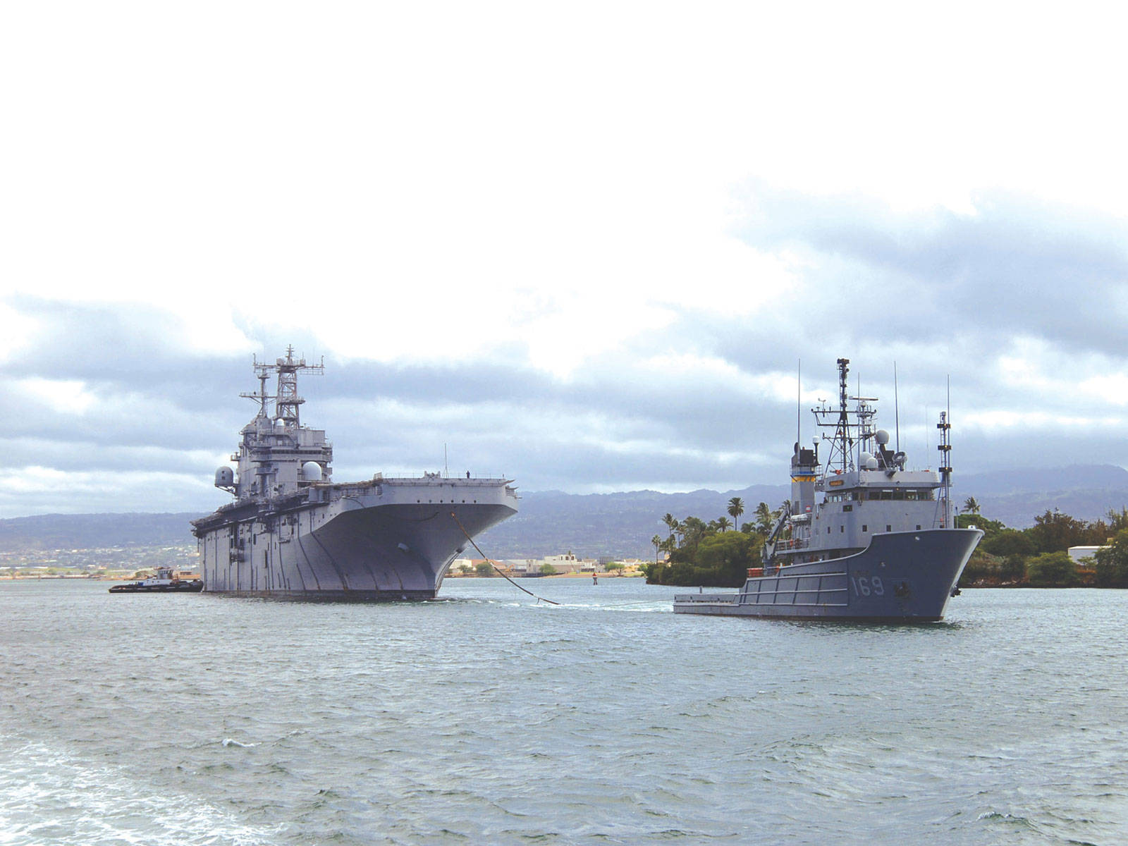 美國海軍艦艇被拖曳 壁紙 — 一艘大型灰色航空母艦和較小的軍艦在陰天、遠山背景下於平靜水域航行。