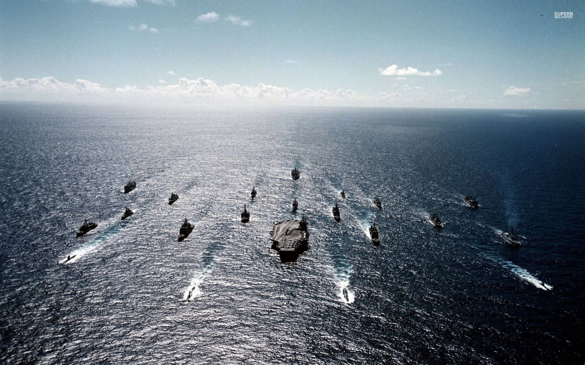 美國海軍艦隊編隊航行 壁紙 — 從空中俯瞰一支海軍艦隊，由航空母艦帶領多艘軍艦，在晴朗天空下穿越廣闊的藍色海洋。
