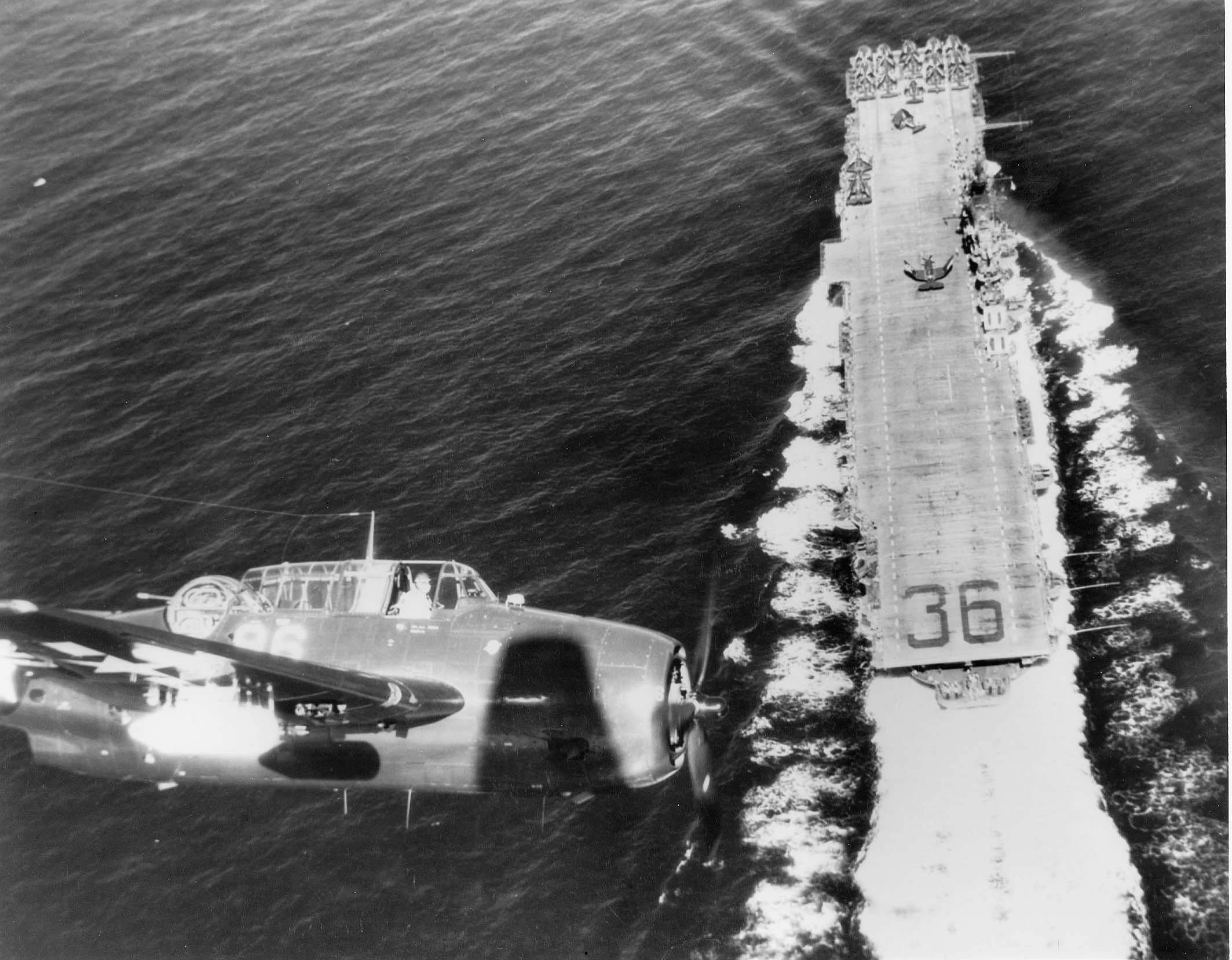 美國海軍飛機飛越航空母艦 壁紙 — 一架復古軍用飛機在一艘航空母艦上方翱翔，航母劃破海浪，呈現戲劇性的黑白航拍畫面。