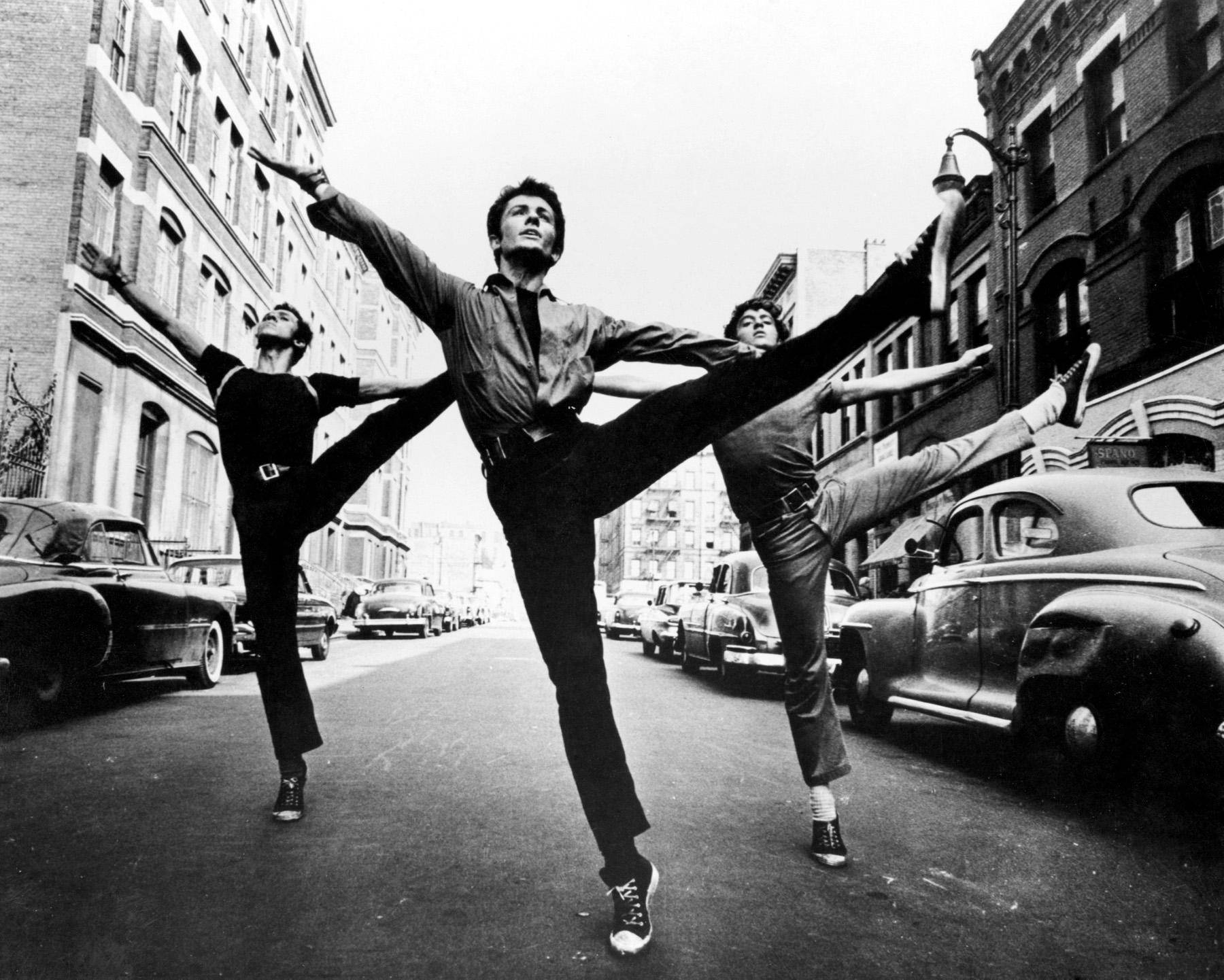 美國演員喬治·查基里斯於1961年《西城故事》中飾演貝納多跳舞的黑白壁紙 壁紙 — 三位舞者在佈滿復古汽車和磚砌建築的城市街道中央擺出戲劇性的芭蕾舞姿勢。