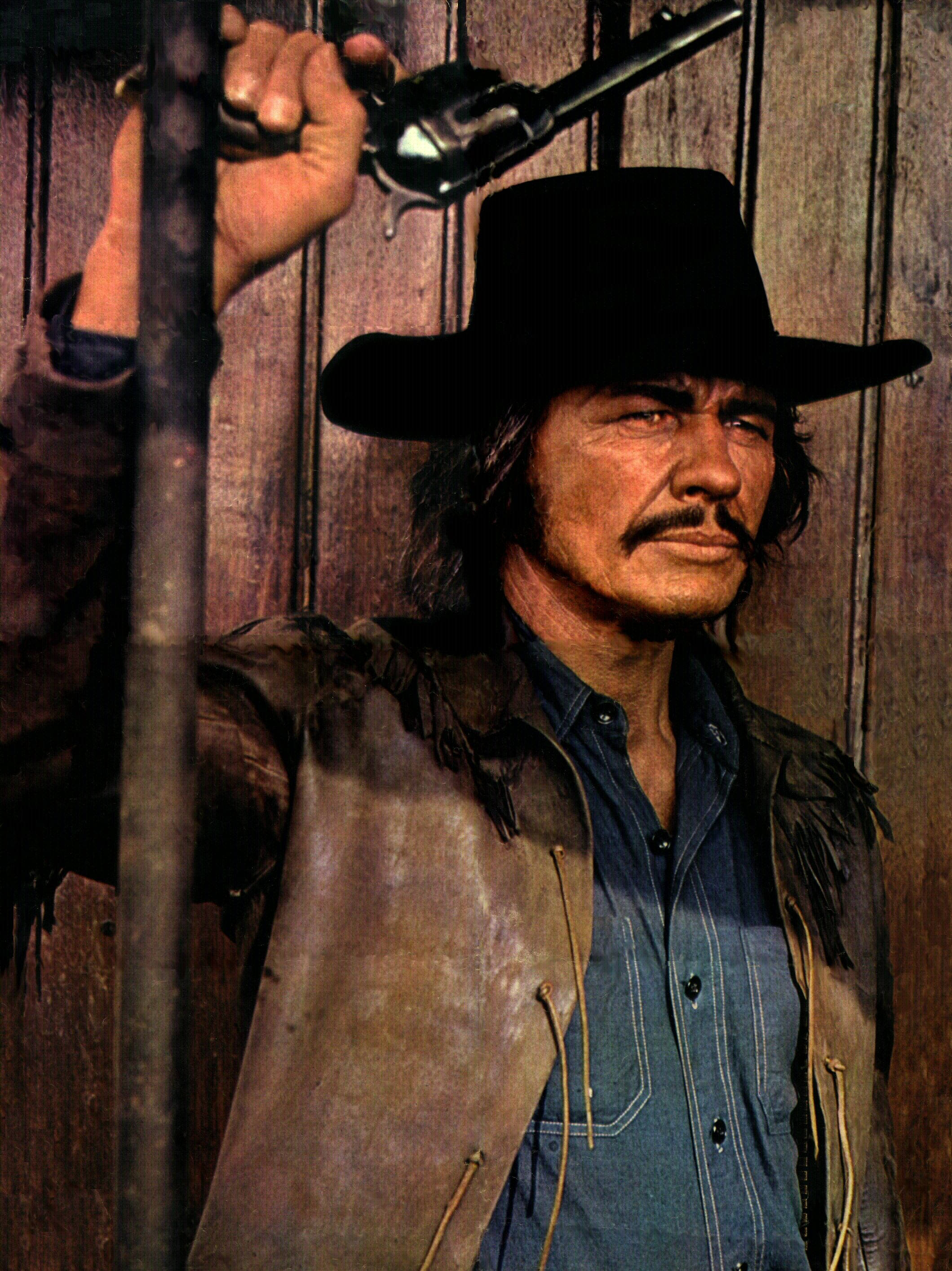美國演員查爾斯·布朗森於1971年電影《紅日》 壁紙 — 一個粗獷的牛仔，留著鬍子，戴著黑帽，手持左輪手槍，對著破舊的木板上，處於緊張的對峙中。
