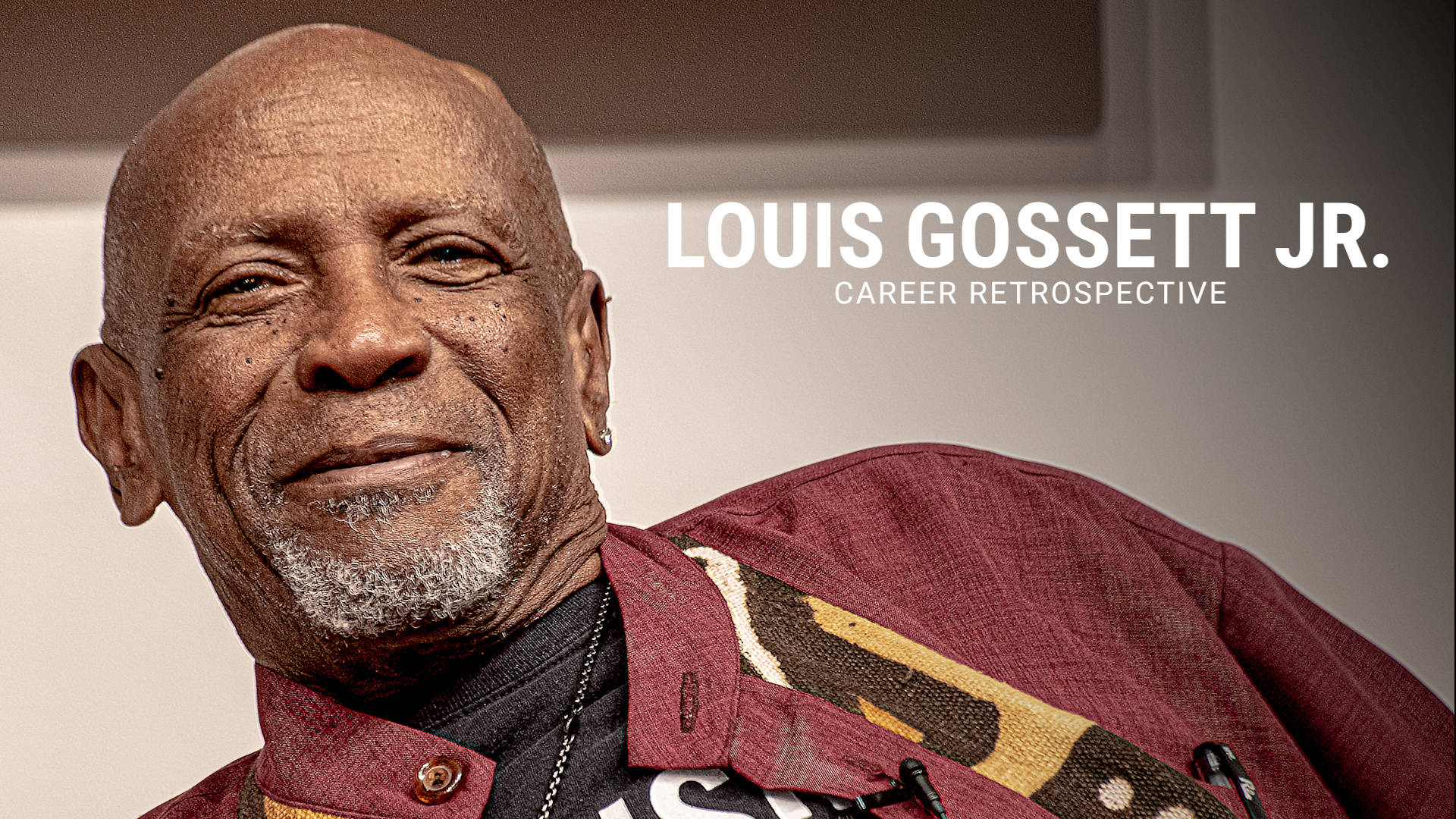 美國演員路易斯·高塞特 Jr. 的職業回顧 壁紙 — 路易斯·高塞特（Louis Gossett Jr.）身穿栗色圖案外套，在中性背景前溫暖微笑的特寫肖像。