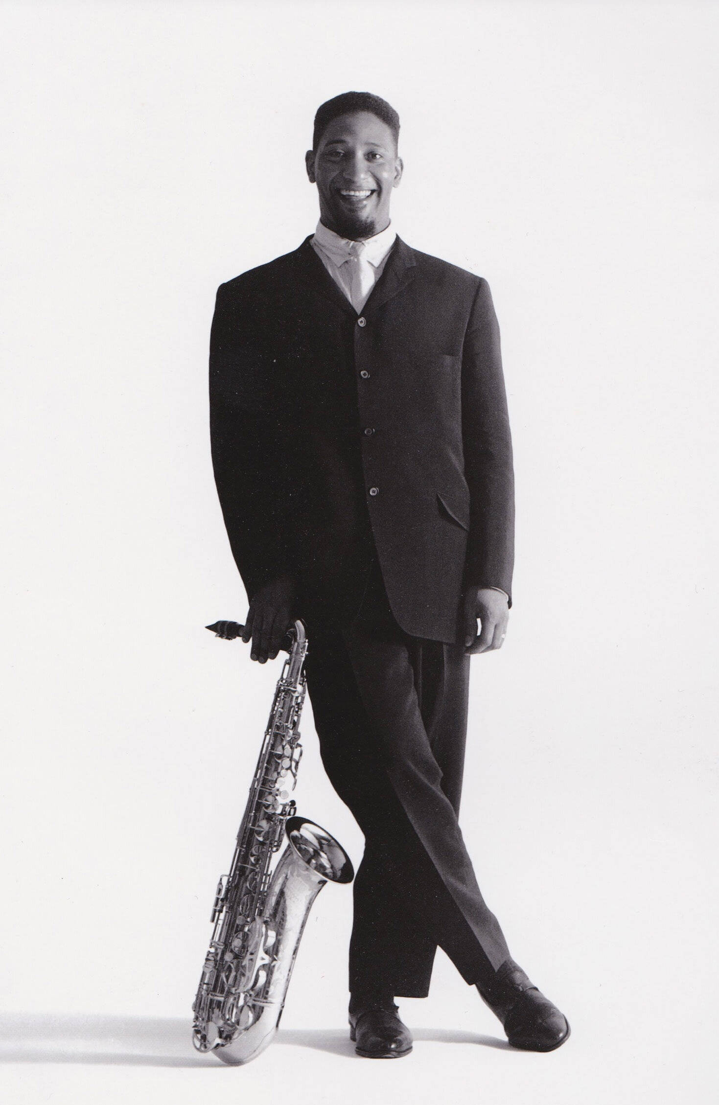 美國爵士傳奇 Sonny Rollins 吹奏薩克斯風 壁紙 — 一個微笑的男人穿著深色西裝，在純白色背景前拿著閃亮的色士風。