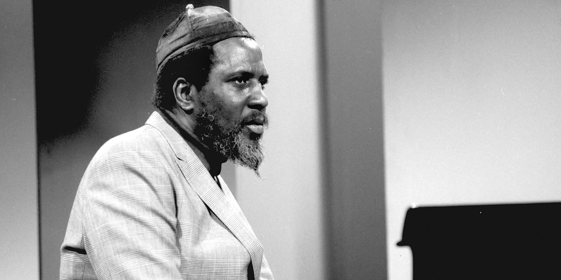 美國爵士鋼琴家Thelonious Monk 壁紙 — 一位戴帽穿西裝外套的鬍鬚男士，在簡約風格的攝影棚中站在鋼琴旁邊的黑白肖像。